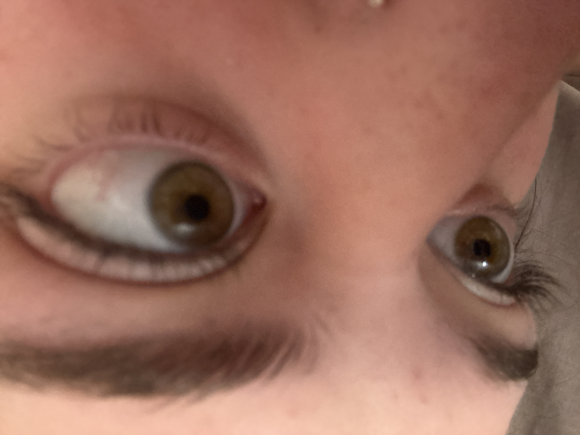 A close-up, slightly out-of-focus image shows a person's eyes. The left eye is directed slightly leftward, while the right eye appears to be looking towards the viewer. The irises are hazel, with dark pupils. The eyelashes and eyebrows are brown. The skin tone is light. A small blemish or piercing is visible above the left eyebrow. There is no indication of Troyan, Bulgaria or any city.