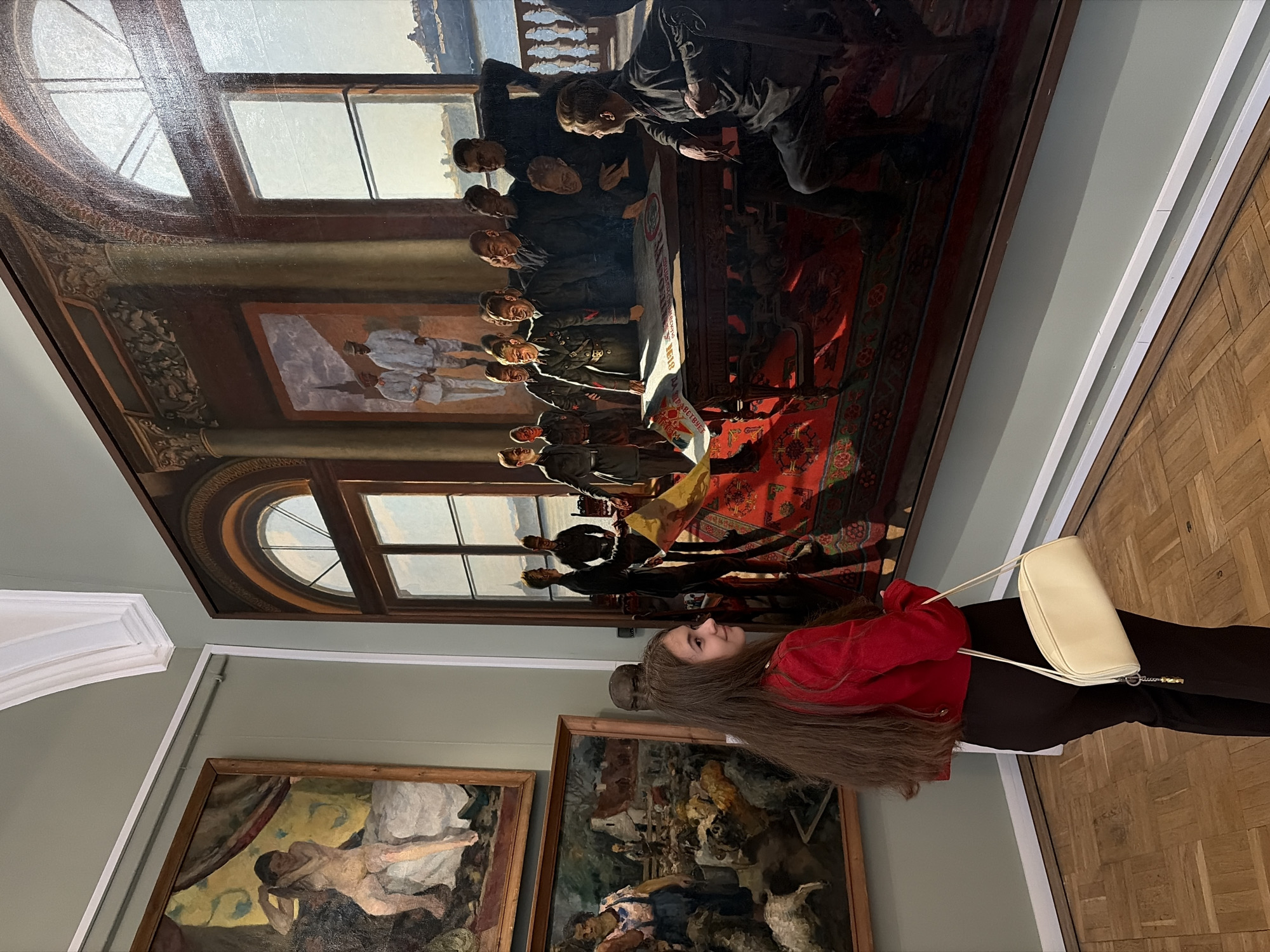 The image shows a young woman in a red jacket and dark pants looking at a large, ornate painting. She is holding a cream-colored purse over her shoulder. The painting depicts a group of men in historical attire gathered around a table. The scene appears to be set indoors, possibly in a museum or gallery, with other paintings visible on the walls. The lighting suggests it is daytime, and the overall atmosphere is one of quiet contemplation. The painting features a red carpet with intricate patterns and a large window in the background. The woman's long brown hair cascades down her back, and she has a thoughtful expression on her face. There is some visible text on a banner within the painting, but it is not legible. The flooring in the foreground is made of polished wood.