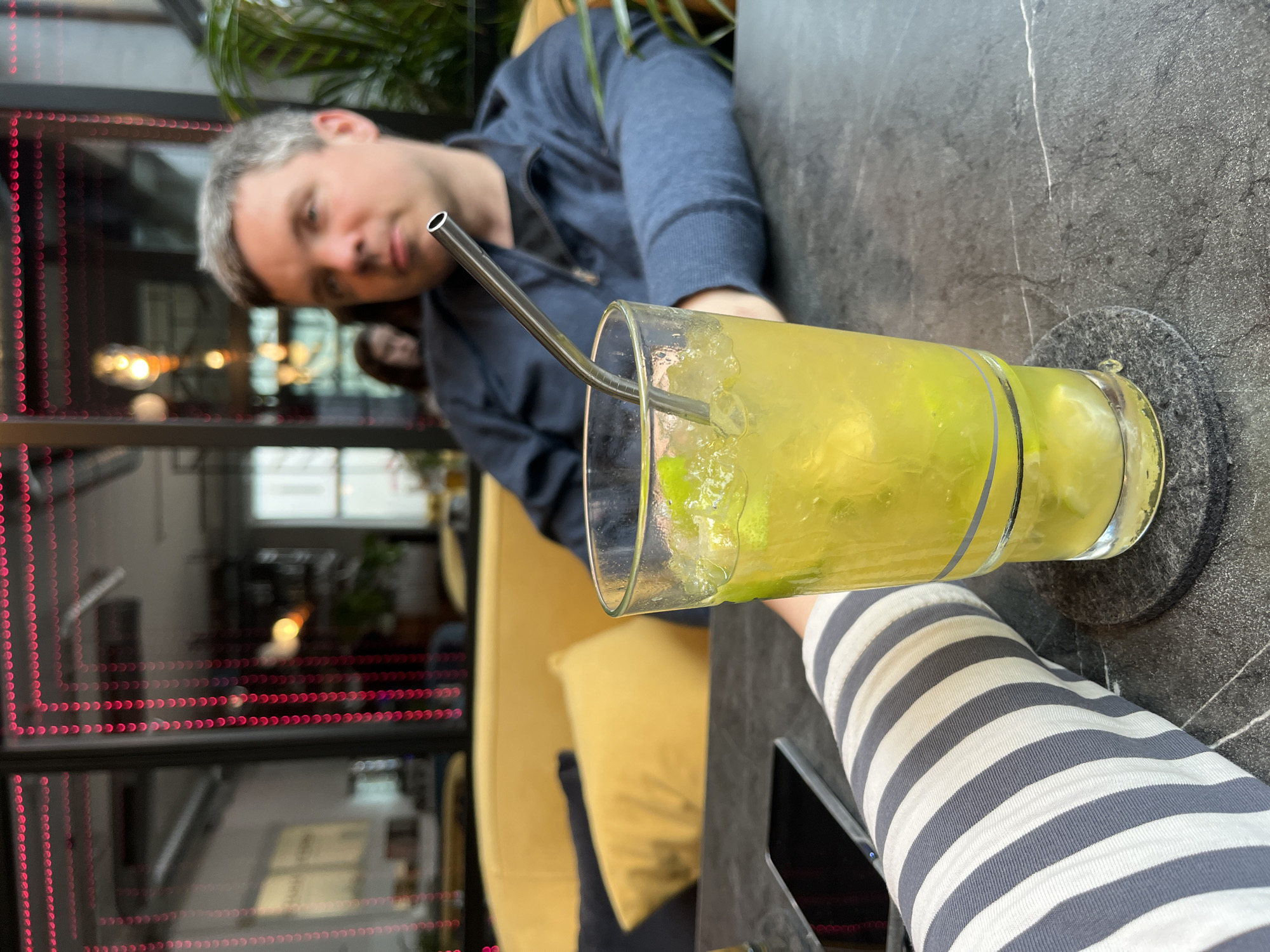 This indoor shot at a café in Bad Kreuznach, Germany shows a man in a blue zip-up jacket seated at a dark gray table, partially obscured by a tall glass of a yellow/green beverage with ice, lime wedges, and a silver straw. The man has graying hair and fair skin. He appears to be looking towards the camera. Seated to the man's right is a person wearing a white and gray striped sleeve. Behind the seated man, a blurred interior of the café is visible, with soft yellow seating and a window illuminated with pink LEDs. A dark grey coaster sits under the glass on the table.