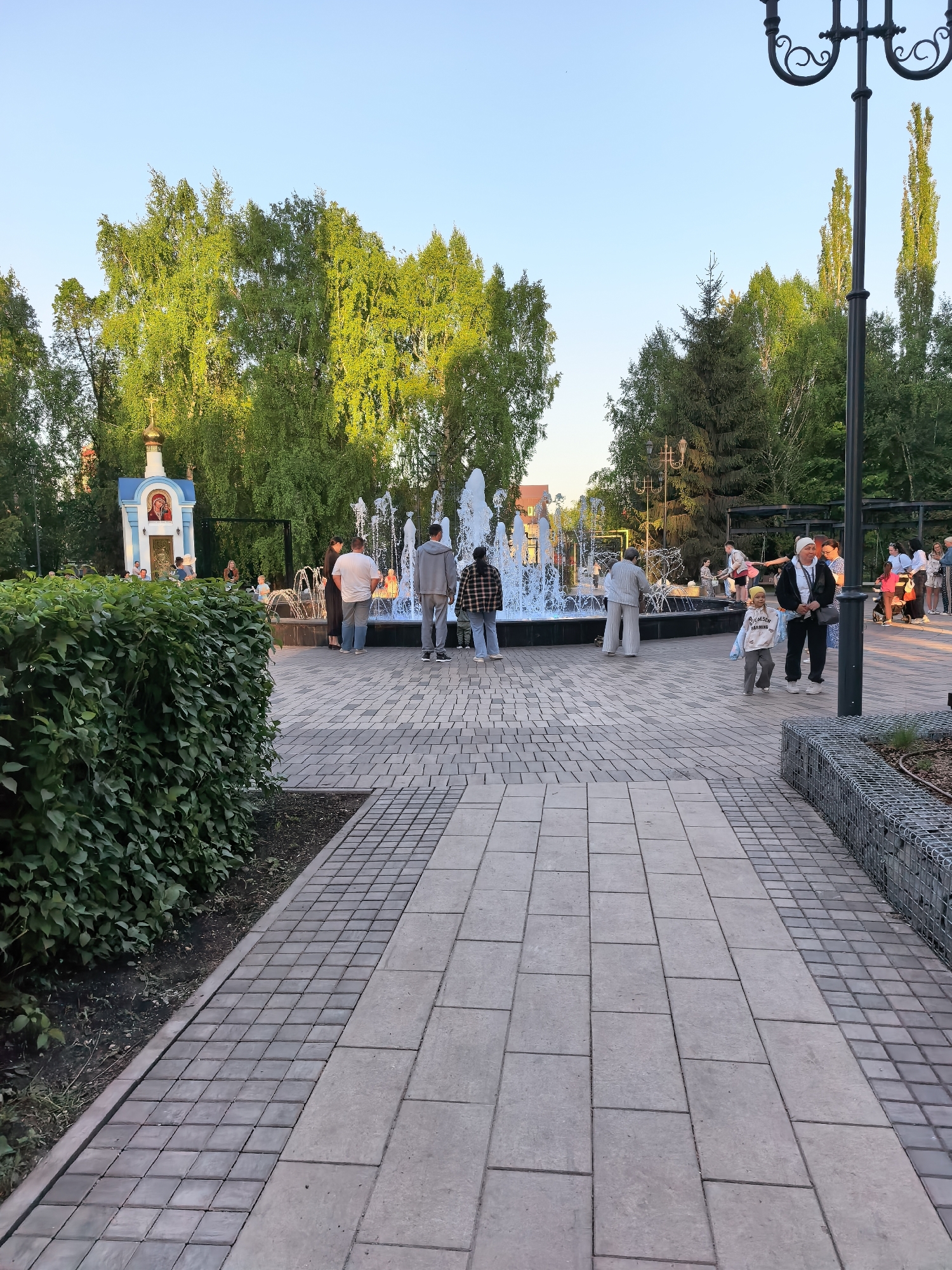The image depicts a park scene in Sterlitamak, Russia, featuring a central fountain surrounded by people. The fountain is actively spraying water. Several individuals are standing near the fountain’s edge, while others are walking or standing nearby. A small, decorated building with an Orthodox Christian icon is visible on the left side of the frame, near a thick green hedge. To the right, a lamppost with ornate metalwork can be seen. The ground is paved with rectangular stones arranged in a patterned layout. A wire-mesh retaining wall borders the walkway on the right. Trees are visible in the background, creating a verdant atmosphere. The sky appears to be a clear, light blue.