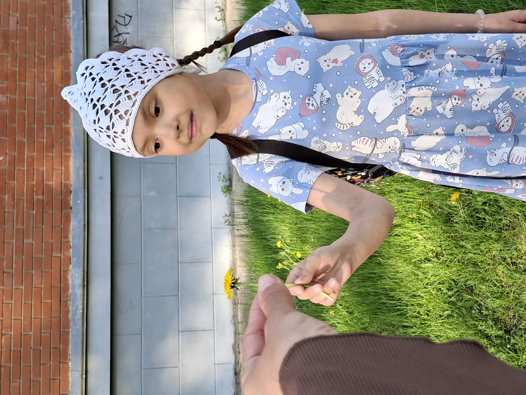 A girl with brown hair in braids, wearing a white crocheted cap and a blue dress patterned with cartoon cats, stands outside in a grassy area. She is near a building with a brick wall on the left and a gray tiled wall next to it. A person wearing a brown garment offers her a yellow dandelion flower. The location is Chaikovsky, Russia.