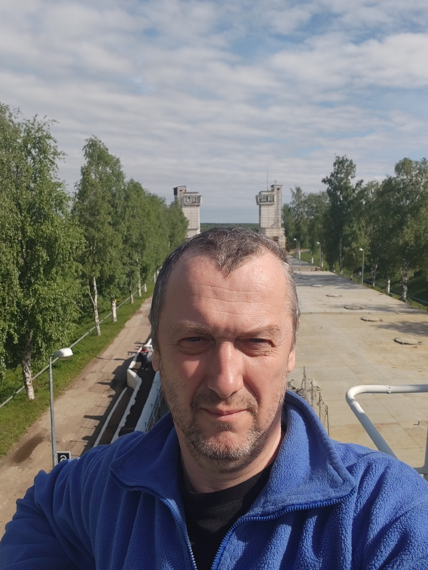 The image presents a selfie of a middle-aged man with short gray hair, wearing a blue fleece jacket over a black shirt. He has a light beard and a serious expression.
The background suggests an outdoor location, possibly on or near a waterway. In the distance, two concrete towers or structures are visible, framed by rows of tall, slender trees lining either side of a concrete pathway or canal embankment. There are regularly spaced lampposts along the path, and the sky is partly cloudy.
The location appears to be a canal or water infrastructure, as seen by the concrete surface in the foreground and the architectural style of the distant towers. The specific city and its location cannot be determined from the visual data.