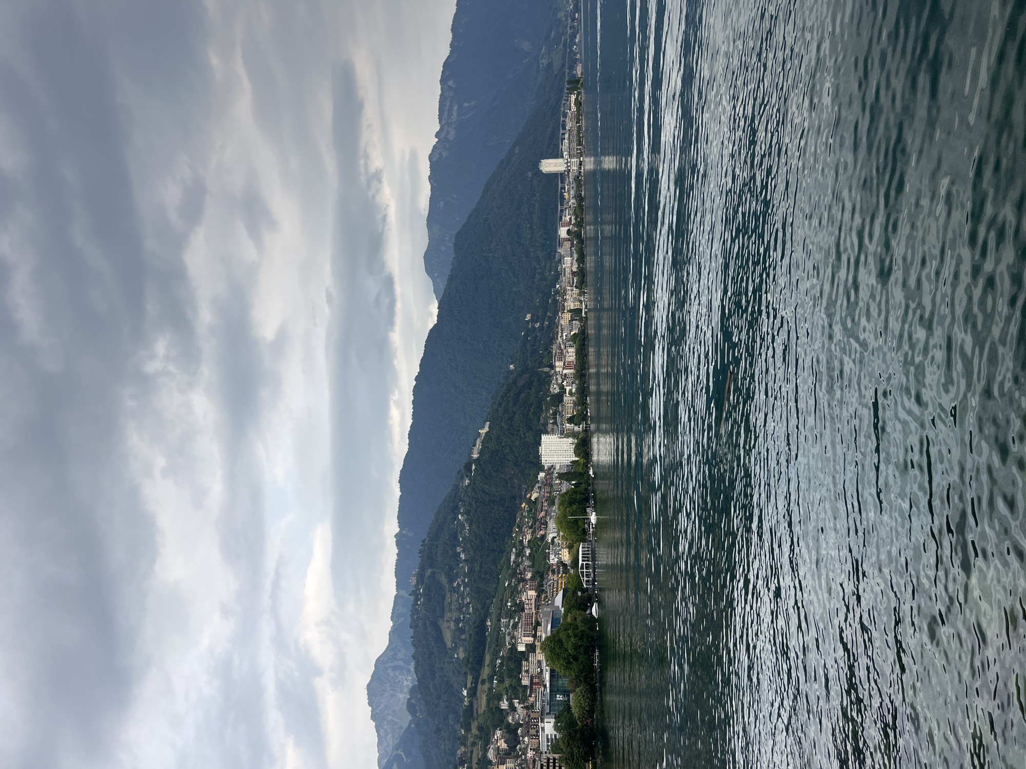 An outdoor, eye-level shot captures a wide, calm body of water in the foreground, with a cityscape and mountains in the background. The water is a dark, choppy blue, with highlights reflecting the overcast sky. The city of Montreux, Switzerland, is nestled along the base of a steep, tree-covered mountain range. Buildings of various heights and architectural styles, ranging from modern white structures to older, more traditional architecture, form a dense urban area. A prominent tall, white building stands out near the center of the city. Green trees dot the landscape, providing a contrast to the buildings and the dark mountains. The sky is filled with heavy, gray clouds, suggesting a slightly gloomy or overcast day. The mountains in the background are imposing, their slopes covered in dense, dark green foliage, with some rocky outcrops visible at higher elevations. The scene appears to be a view from a boat on Lake Geneva, looking towards the Montreux shoreline.