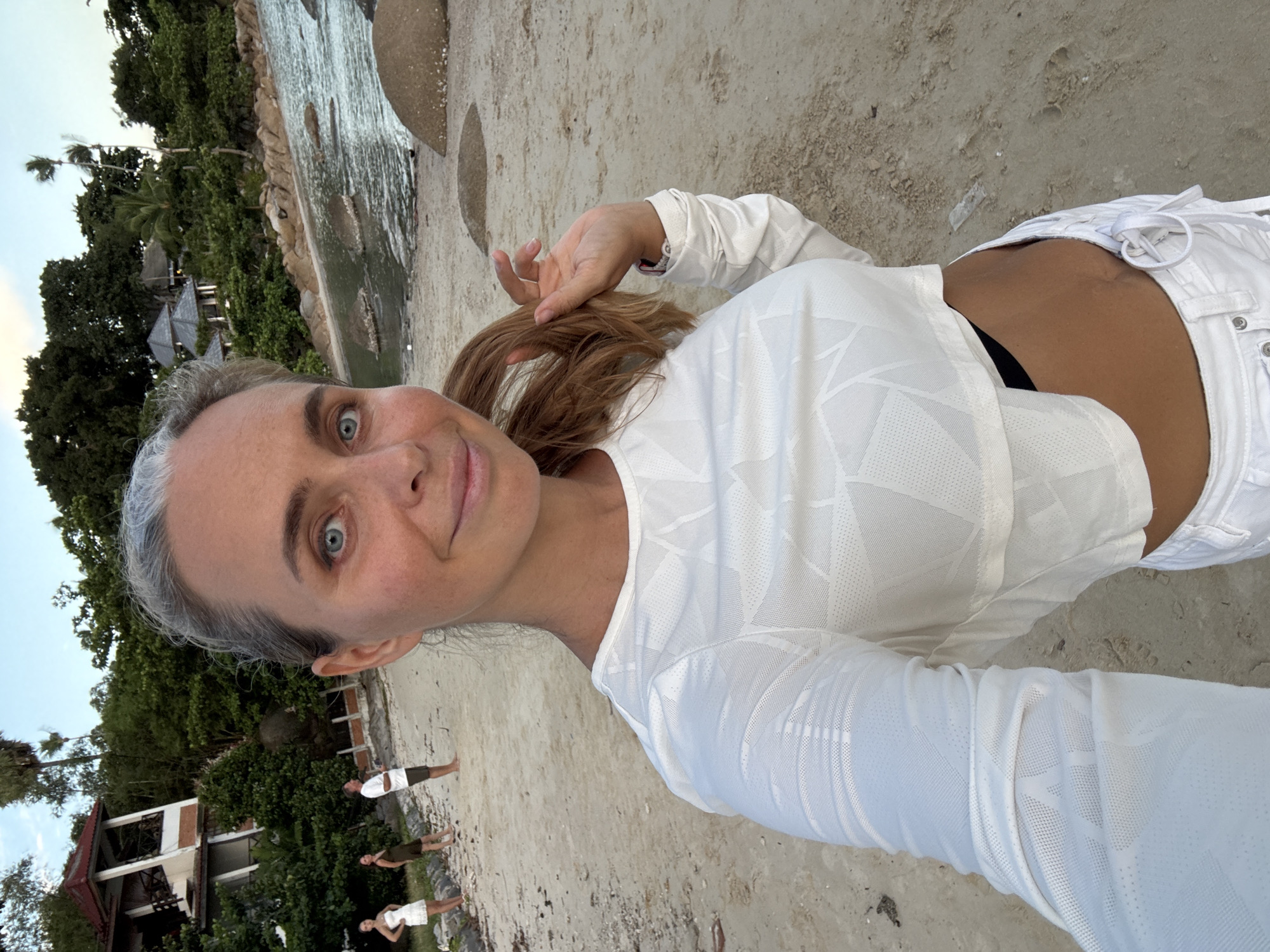 A woman with graying blonde hair and blue eyes is taking a selfie on a sandy beach in Ko Samui District, Thailand. She is wearing a white cropped long-sleeved shirt with a geometric pattern and white shorts. Her left hand is touching her hair. In the background, two other women and a man are standing on the beach. Behind them are lush green trees, rocky formations, and buildings. The ocean is visible to the right of the image, with rocks in the water. The sky is overcast.
