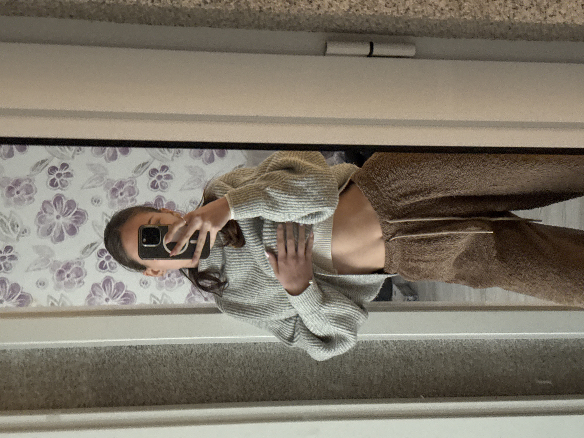 An indoor, vertical mirror selfie captures a person standing, looking down at a smartphone held up to their face. The image is originally rotated 90 degrees counter-clockwise. The individual, appearing to be female, wears a light gray, long-sleeved, ribbed cropped sweater that exposes her midriff, paired with brown, textured, high-waisted sweatpants with visible drawstrings. One hand holds the black smartphone, partially obscuring her face, while the other hand rests gently on her upper stomach. Rings are visible on her fingers.
The background visible in the mirror reflects a room with a white and purple floral wallpaper pattern behind the person's head and upper body. Below this, a white frame (possibly a door or mirror frame) separates the wallpaper from a darker, textured surface, likely a carpeted floor or lower wall. Above the mirror's reflection, a similar textured surface, perhaps a ceiling or upper wall, is visible. The lighting appears to be indoor, and no specific time of day or weather can be discerned. There is no visible text in the image. The scene is located in Sofia, Bulgaria.