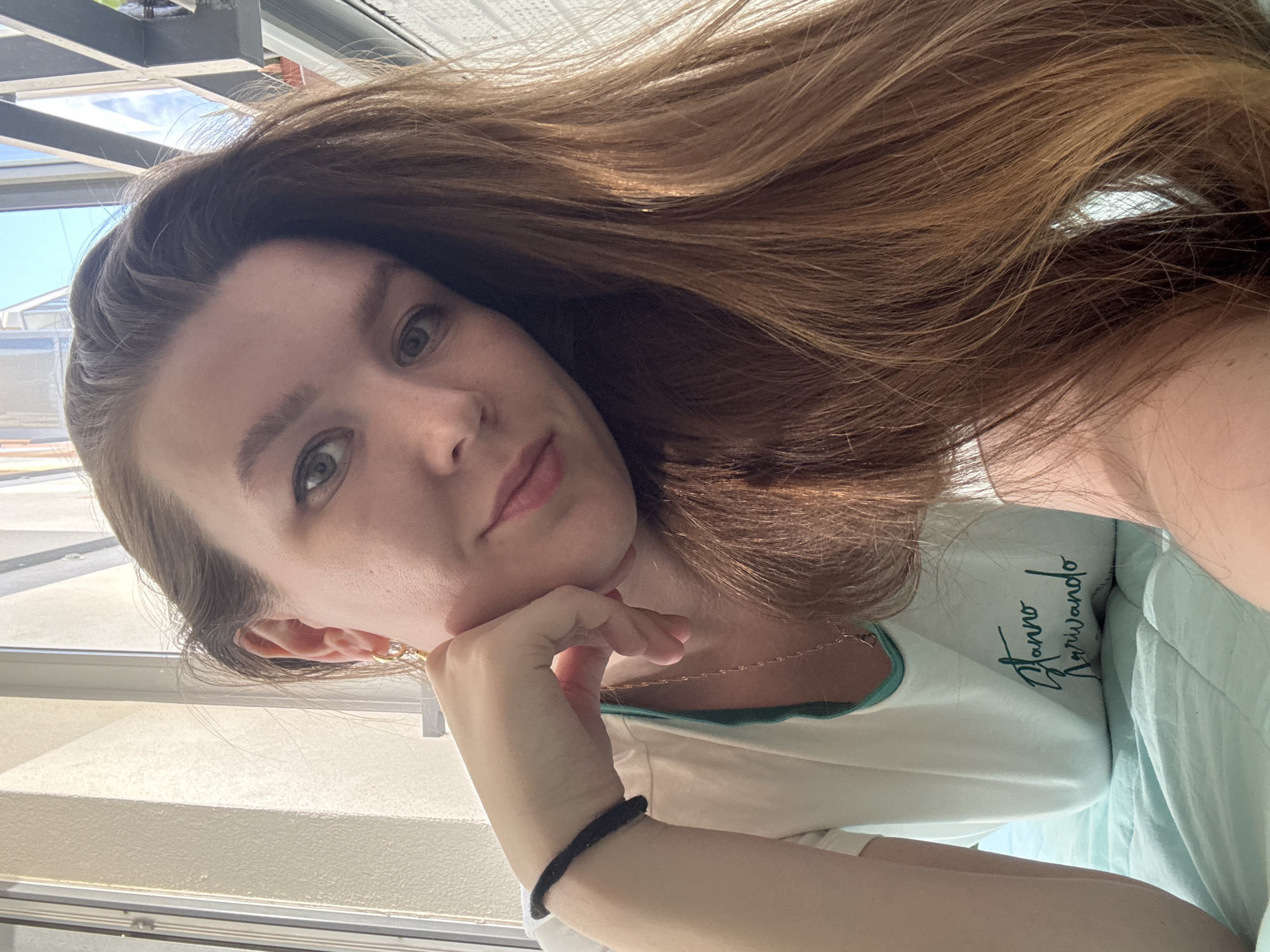 A woman with long brown hair is posing for a selfie in front of a window. She is wearing a white t-shirt with green trim and the word "Starro Variwando" printed on it. She has her hand on her chin and is looking at the camera. The background is blurry, but it appears to be a city scene with buildings and sky visible through the window. The image does not show anything that identifies the location as Izhevsk, Russia.