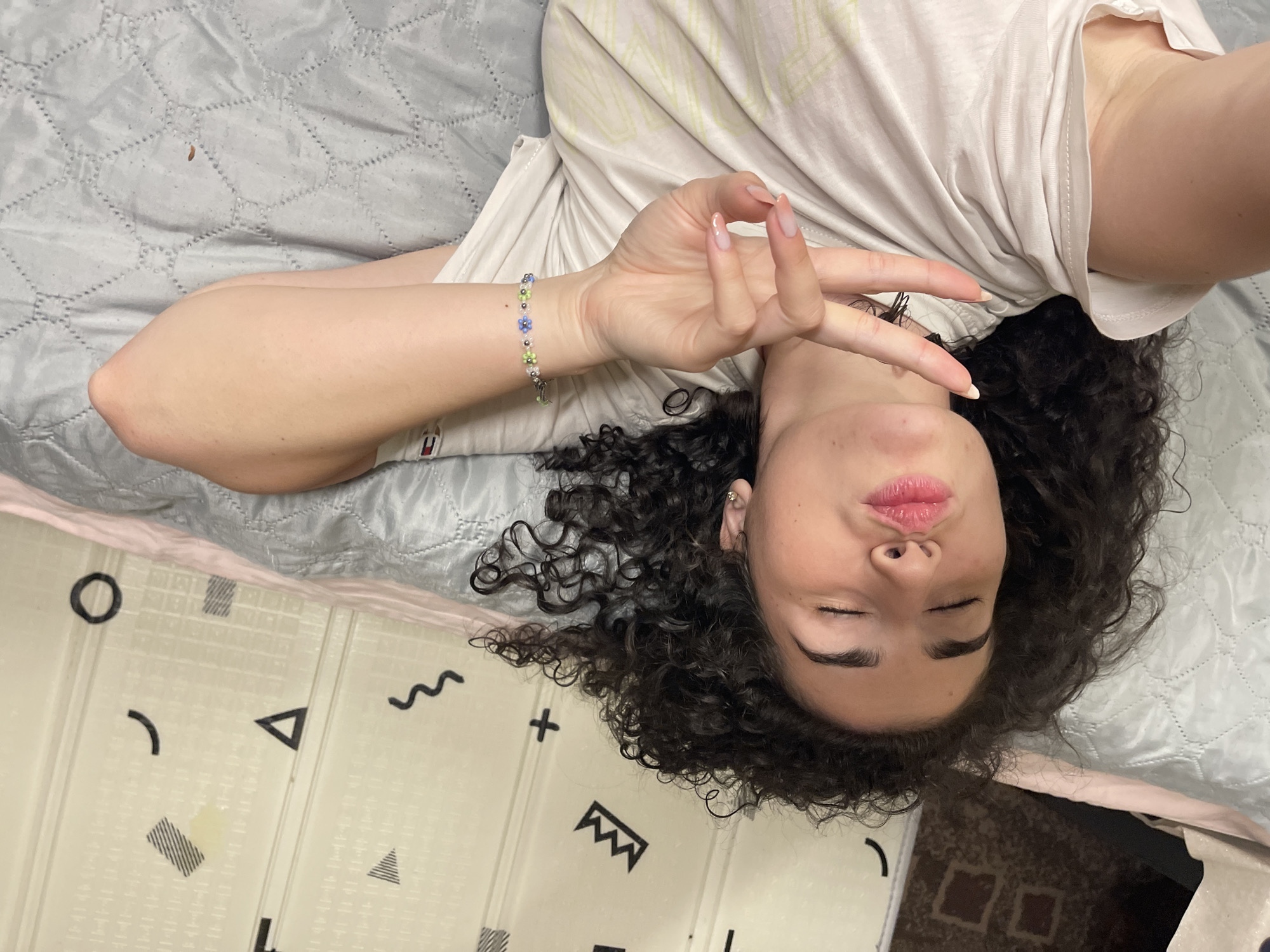 The image depicts a young woman with curly dark hair lying on her side on a quilted surface, possibly a bed or a sofa. She is wearing a light-colored t-shirt. Her eyes are closed, and her lips are puckered as if blowing a kiss. Her right hand is raised, forming a peace sign with her index and middle fingers, while her thumb and ring finger are touching, creating a circle. She is also wearing a beaded bracelet on her wrist. The background consists of a surface with a geometric pattern, resembling a dresser or a wall. The overall lighting is soft and natural. The image does not provide any specific visual cues that would allow for pinpointing a location within the city of Sofia, Bulgaria.