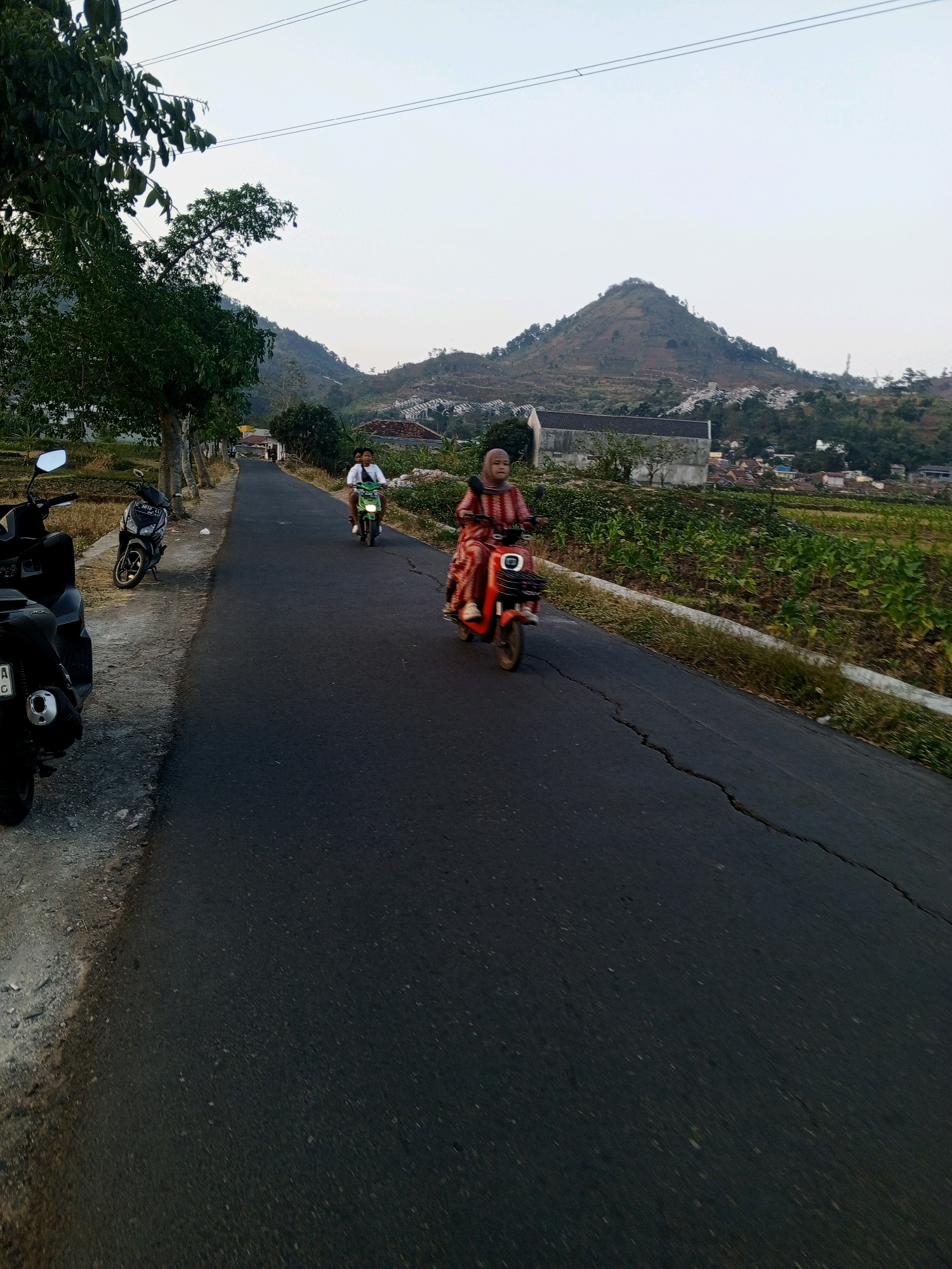 In an outdoor, rural setting, a paved road curves through a landscape of rolling hills and vegetation under a clear sky. Trees line the left side of the road, with several parked motorcycles visible. Two people are riding motorcycles on the road. A person in a pink and orange patterned outfit on a red electric scooter is in the foreground, moving away from the viewer. Further up the road, a person in a white shirt is on a green motorcycle, also moving away. In the background, a large, conical hill dominates the horizon, with scattered houses and terraced fields on its slopes. The road surface is dark asphalt with visible cracks. The scene is captured from a slightly elevated perspective, looking down the road.