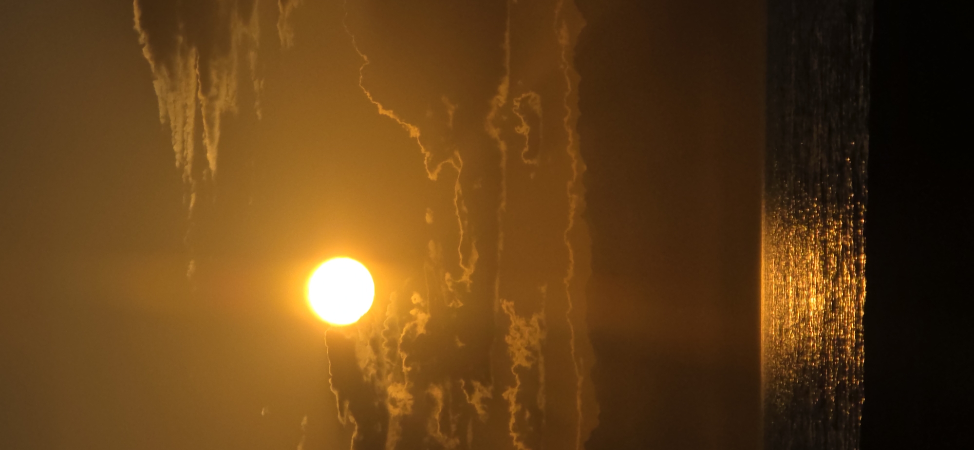 A photograph captures a sunset over a body of water, likely a sea or large lake, with a dark foreground suggesting a beach or land. The sun, a brilliant white orb, is positioned in the upper left quadrant, partially obscured by wispy, golden clouds. Rays of light fan out from the sun, creating a dramatic atmospheric effect. The water reflects the intense golden light of the sunset, creating a shimmering, almost sparkling surface. The horizon line is clearly defined, separating the water from the hazy sky. The overall color palette is dominated by warm oranges and yellows, indicative of a sunset. The image does not contain any people or specific urban landmarks that would identify it as being in Basbeshik, Kyrgyzstan.