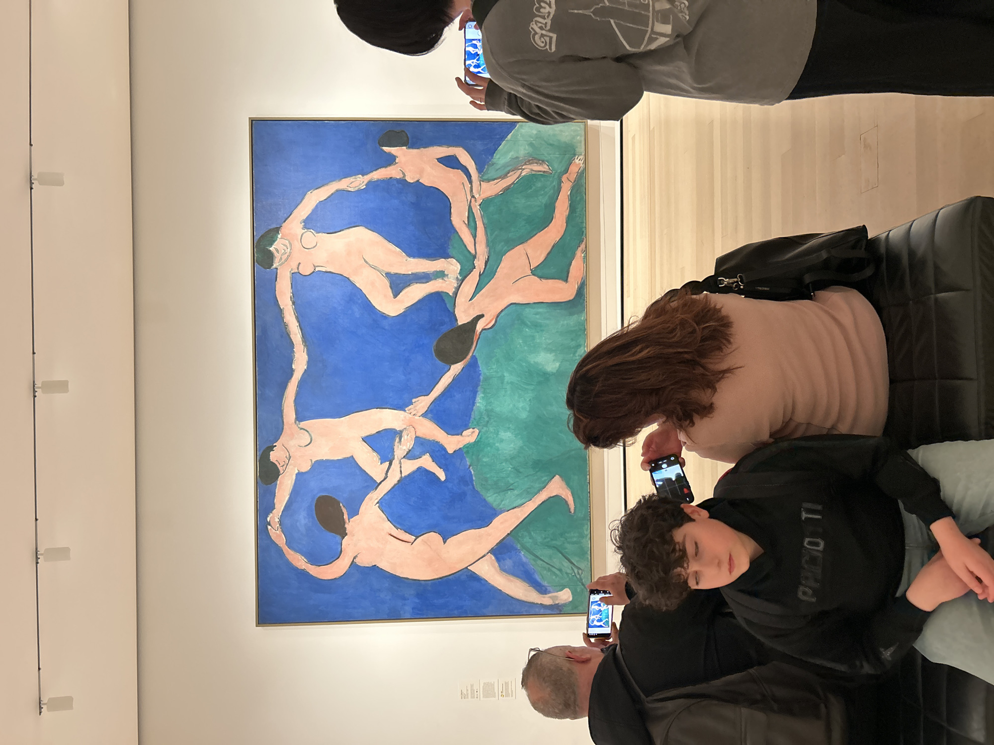 The image depicts a group of people viewing Henri Matisse's painting "Dance" at the Museum of Modern Art (MoMA) in New York City. The painting shows five nude figures dancing in a circle against a blue and green background. Three individuals are actively taking pictures of the artwork using their smartphones, while two others are seated on a bench. One of the seated individuals is resting with their eyes closed. The environment appears to be a well-lit gallery space.