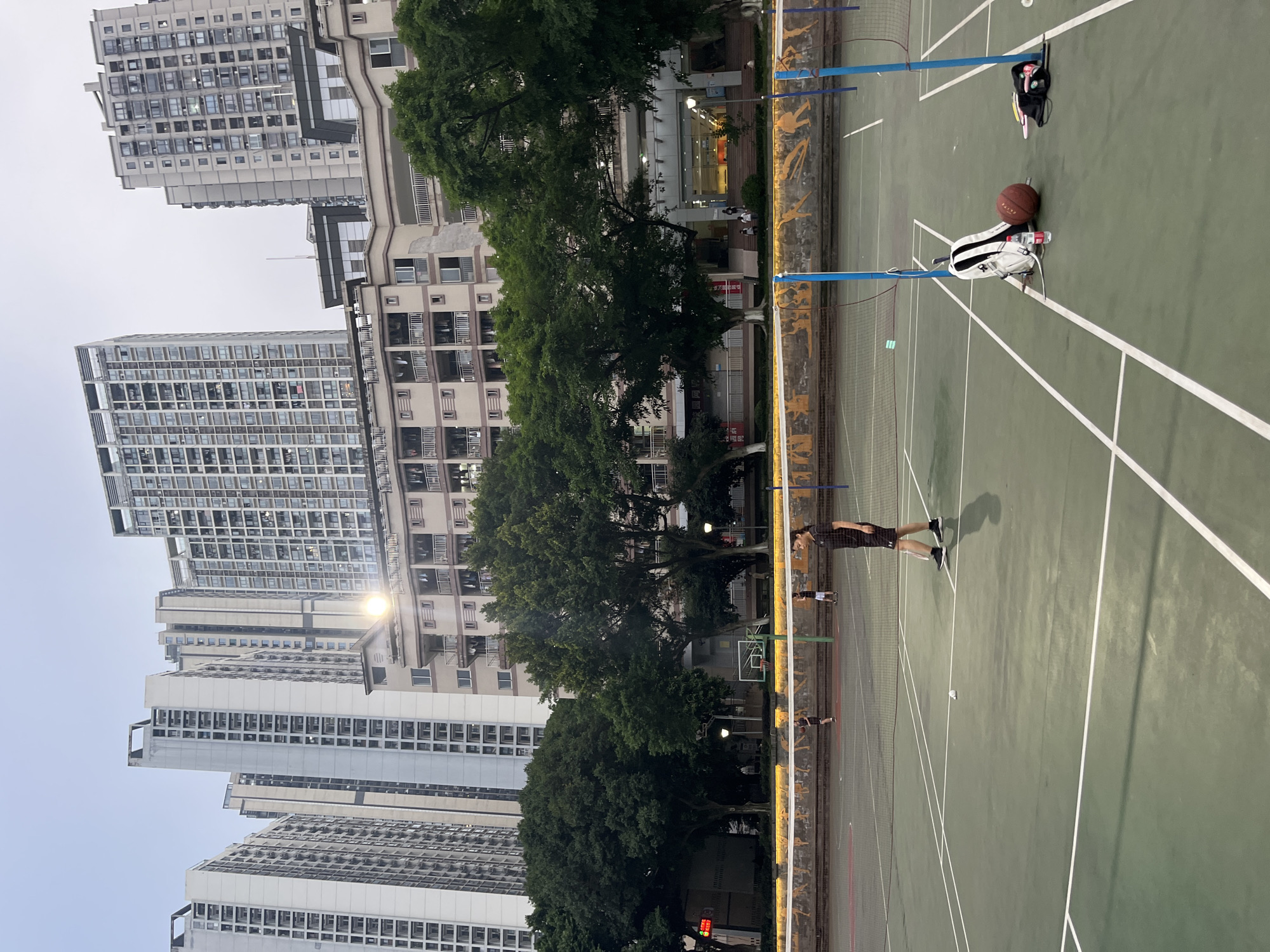 The image depicts an outdoor badminton court in the Yuzhong District of China, with tall residential buildings looming in the background. The court surface is green with white markings. A net is strung across the court. In the foreground, on the right side of the court, a white backpack, a basketball, a water bottle, and a black bag with pink accessories are placed on the ground. A person wearing a black t-shirt and shorts is standing on the court, holding a badminton racket. Two other individuals are visible on the court, engaged in playing badminton. The surrounding environment includes trees and a decorative wall with murals along the edge of the court. The sky is overcast, indicating a twilight or cloudy day.