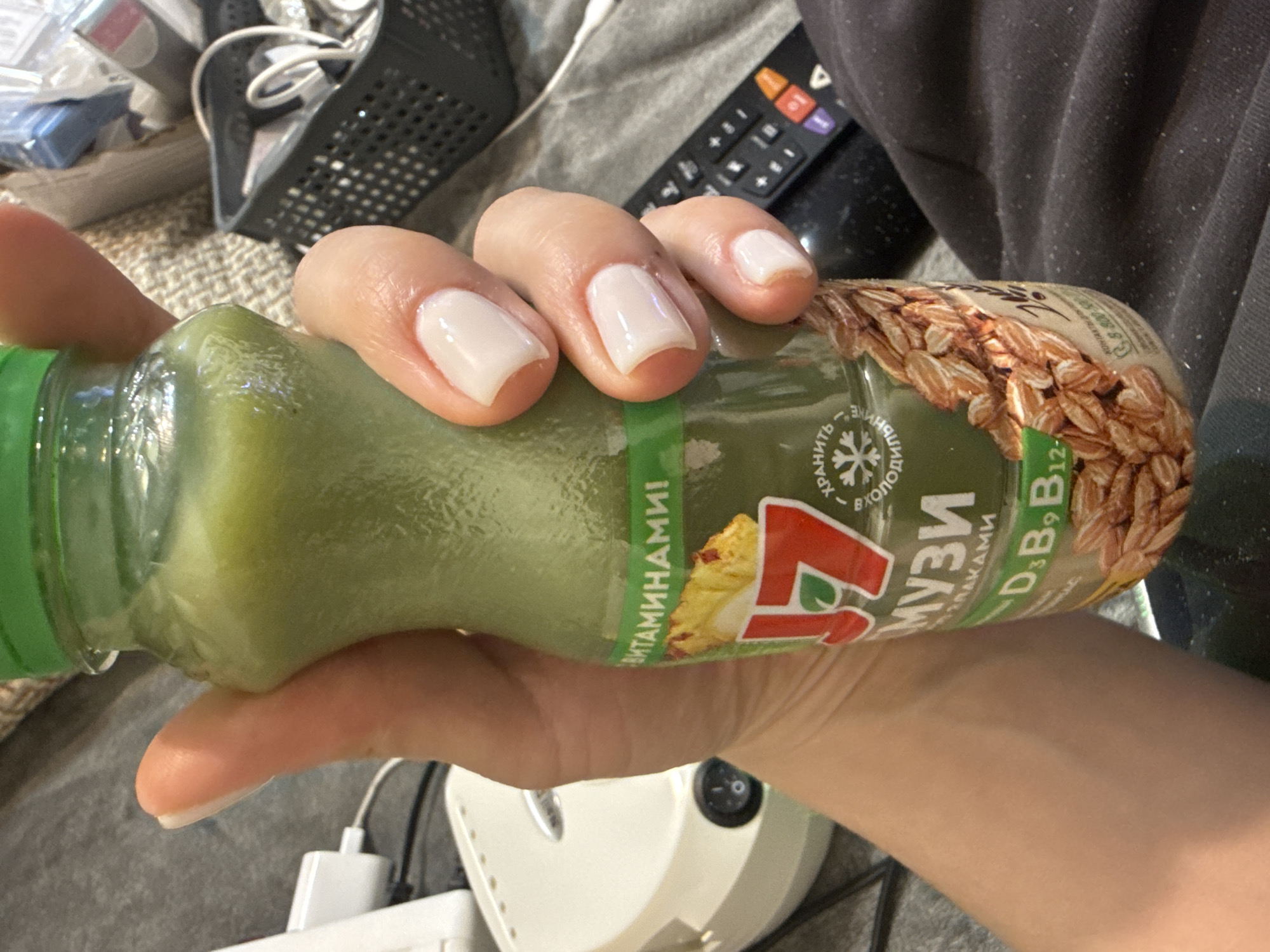 The image depicts a person holding a bottle of what appears to be a green smoothie or juice. The bottle has a green cap and a label in Russian that reads "Музи" (Muzi), "с витаминами!" (with vitamins!), and lists vitamins D3, B9, and B12. The label also features an image of what looks like pineapple and an illustration of oats.
The person's hand is visible, with light-colored nail polish. In the background, there is a television remote, a black plastic container, and a white humidifier.
Based on the Russian labeling on the product, the location might be Russia. However, without additional information, pinpointing the exact location in Mytishchi, Russia, is not possible.