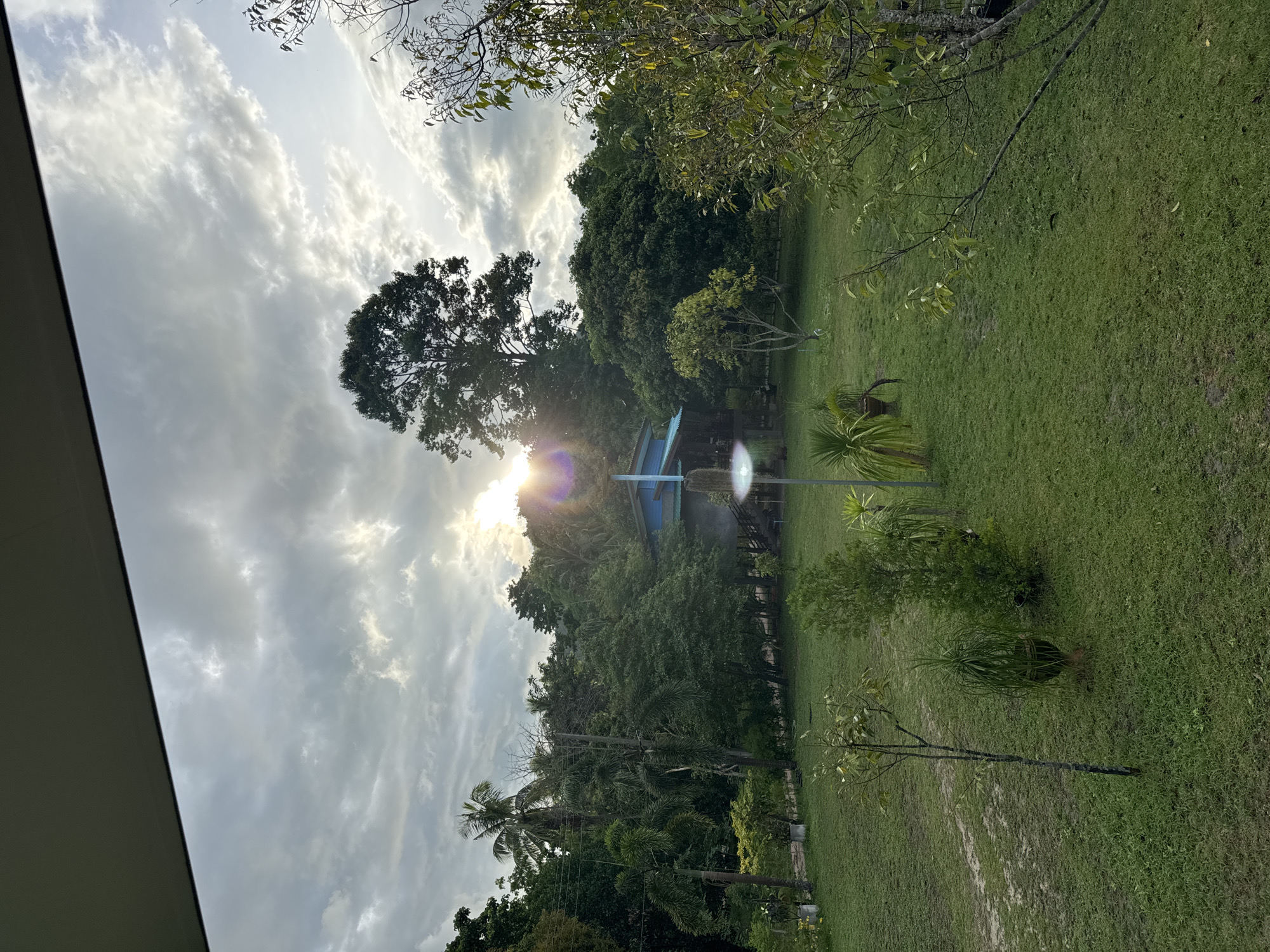 The image depicts an outdoor scene, likely a residential or park area in Baan Don Sai, Thailand. The landscape consists of a grassy lawn bordered by various trees and shrubs, suggesting a natural or landscaped environment. In the distance, a building with a blue roof is partially visible through the trees. Overhead, the sky is filled with dramatic, textured clouds and the sun is shining brightly through, creating a lens flare effect. The image appears to have been taken from a slightly elevated position, possibly from within a building or structure, as a portion of a roof or wall is visible in the lower left corner. There are no people visible in the image.