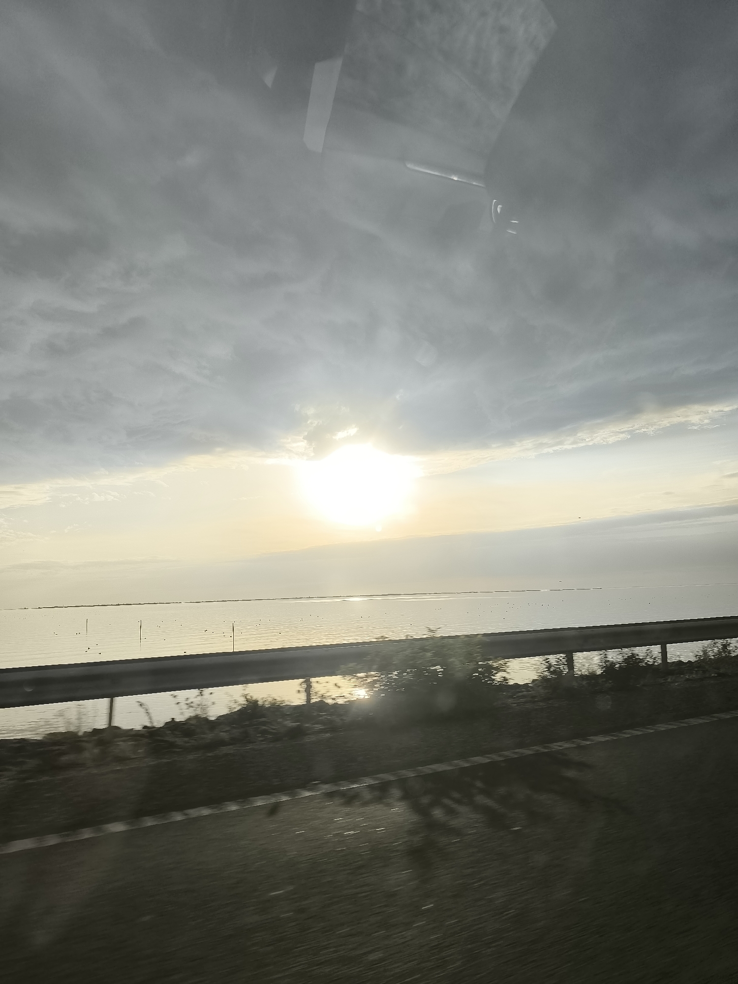 The image captures a view of a body of water, possibly a lake or sea, under a cloudy sky with the sun partially obscured by clouds. The sun is a bright, diffused light source, casting a soft glow on the water's surface. There are some small, dark objects scattered across the water, which could be birds or buoys. A metal guardrail runs horizontally across the middle ground, separating the water from a road or path in the foreground. The foreground also shows a blurred asphalt surface with some reflections, suggesting movement or a wet surface. In the upper portion of the image, reflections of a vehicle's interior, including part of the dashboard and windshield, are visible, indicating the photo was taken from inside a moving vehicle. The scene appears to be a roadside view, likely along a highway or coastal road. The overall impression is one of a somewhat overcast day with a glimmer of sunlight breaking through. Based on the context provided about Lelystad, Netherlands, this could be a view along the Houtribdijk, which separates the IJsselmeer and the Markermeer, or a similar stretch of water accessible by road in the area.