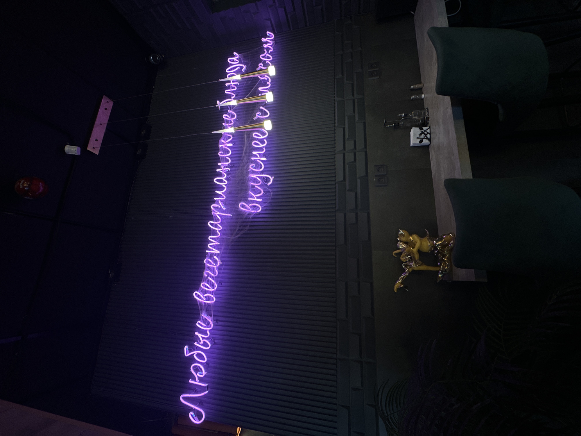 The scene depicts the interior of a dimly lit establishment, likely a restaurant or bar, located in Rostov-on-Don, Russia. A prominent purple neon sign on the upper wall displays the Cyrillic text "Любые вегетарианские блюда вкуснее с мясом," translating to "Any vegetarian dishes are tastier with meat." The wall is clad with dark grey vertical ribbed panels in the upper section and horizontal geometric panels below. The dark ceiling features a red fire alarm device, a white cylindrical sensor, and three gold-capped pendant lights suspended from a wooden base. A dark circular vent is visible on the upper right ceiling. Below the neon sign, a light-colored counter extends horizontally, accompanied by two dark-upholstered bar stools with backs. Items on the counter include a golden figurine depicting two stylized figures, a white napkin dispenser with a cutlery logo, a clear glass bottle, and two shaker-style containers. Dark foliage is present in the lower-left foreground. No individuals are visible.