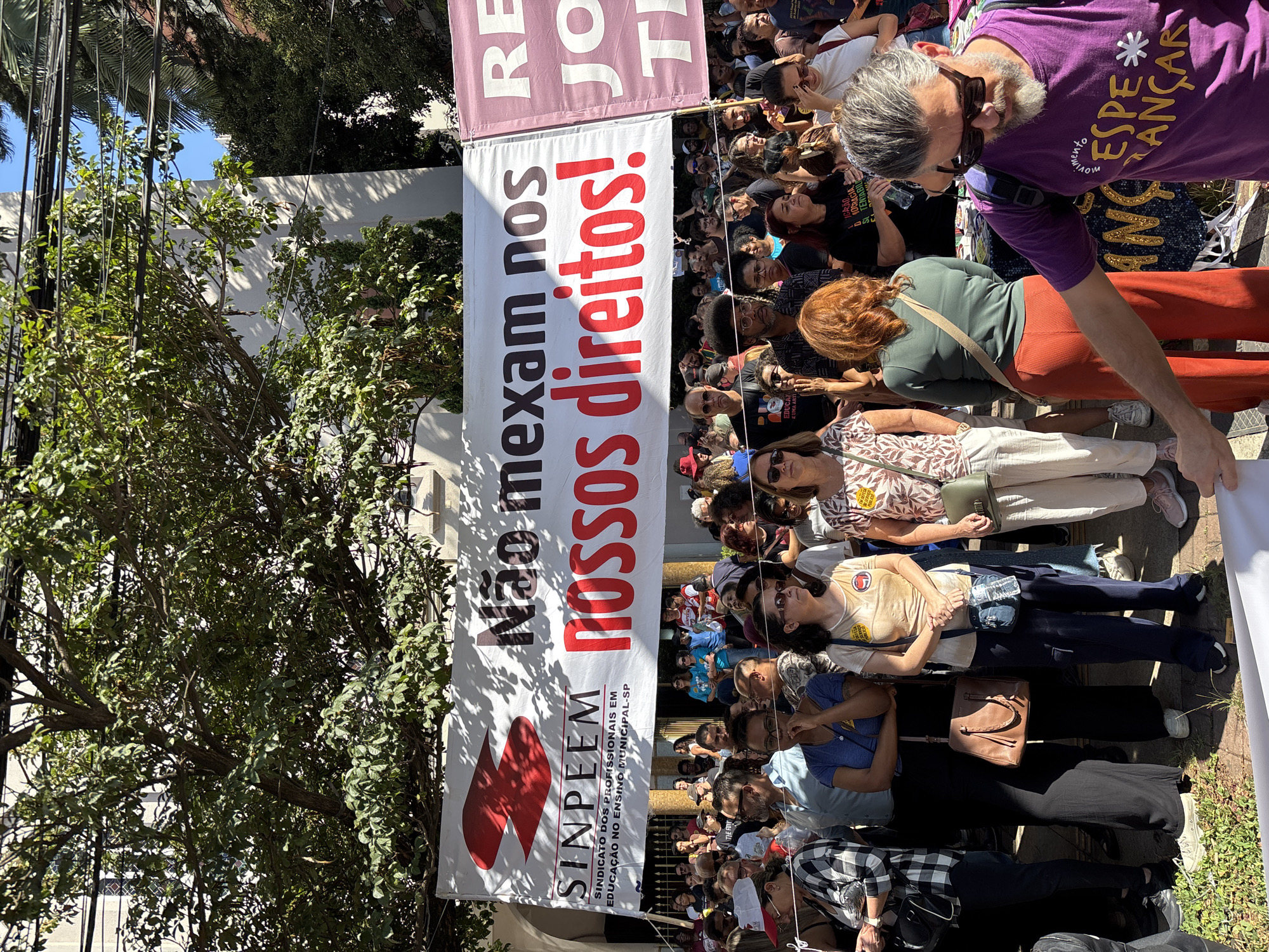 A high-angle, outdoor shot shows a large group of people participating in a demonstration or protest, possibly in the city of São Paulo, Brazil. The crowd is dense, composed of individuals of varying ages and appearances. Many are looking in the same direction, likely towards a focal point of the demonstration.
The demonstrators are holding a large banner with a message in Portuguese: "Não mexam nos nossos direitos!" which translates to "Don't mess with our rights!" Below this is the text "SINPEEM SINDICATO DOS PROFISSIONAIS EM EDUCAÇÃO NO ENSINO MUNICIPAL-SP," indicating that the participants are likely teachers or educational professionals.
The setting appears to be a street lined with trees, and a building is visible in the background, partially obscured by the banner and trees. Sunlight suggests it is daytime. People are dressed in casual attire. The photo is taken from above the crowd, providing a wide view of the event.