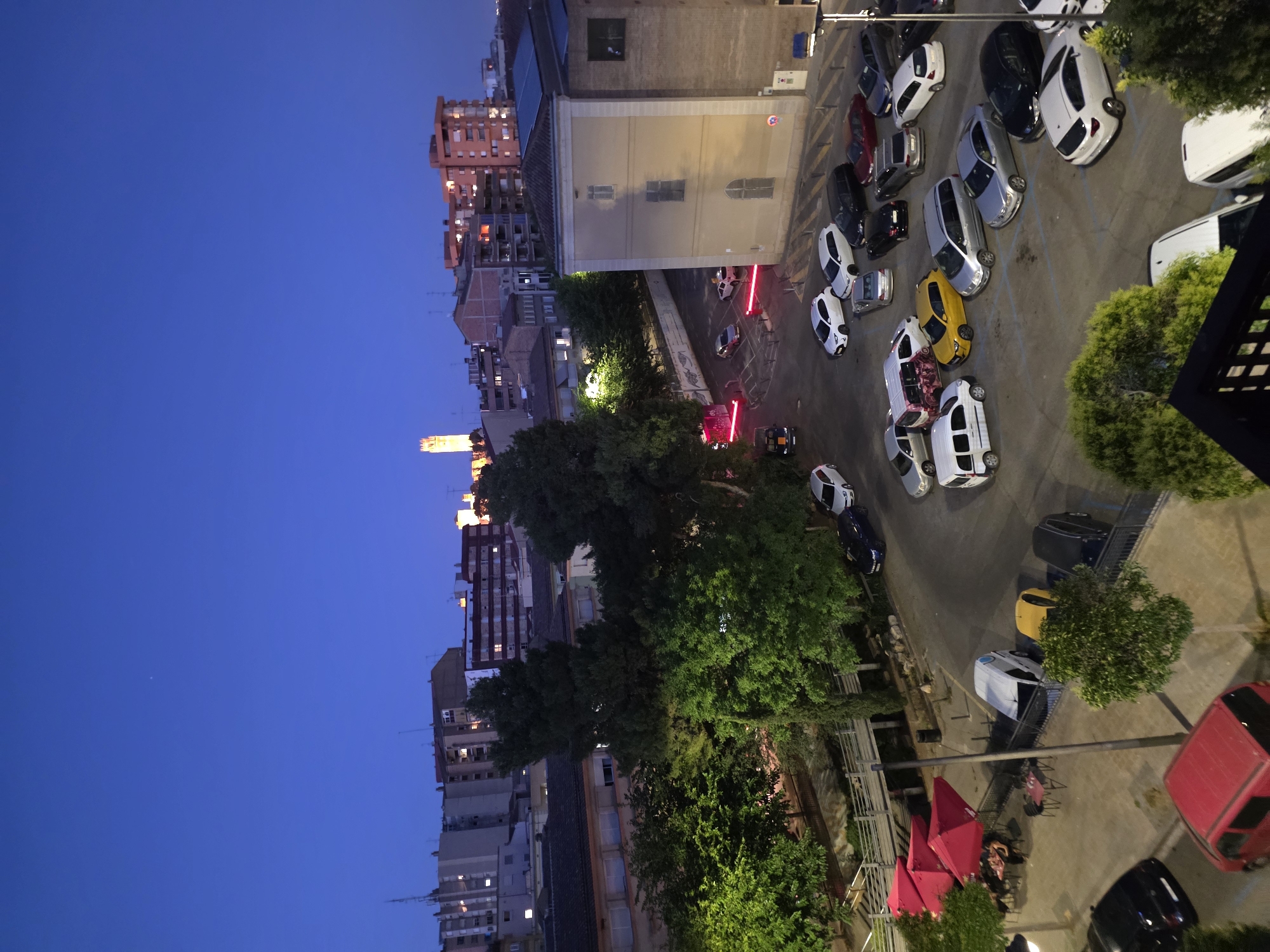 An aerial, rotated view looking south at the city of Lleida, Spain, at dusk or twilight. The sky is a deep blue. On the left are tall buildings with lit windows. Trees are visible in the middle of the image. On the right is a parking area with many cars, including white, yellow, red, and black vehicles. There's a building to the upper right of the parking lot. Additionally, there's an outdoor dining area with pink umbrellas and people seated at tables.