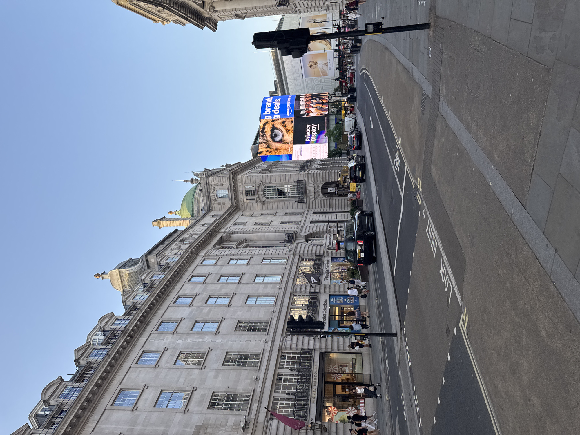 The image captures a street scene in London, United Kingdom, likely Piccadilly Circus, given the iconic architecture and illuminated billboards. The photograph is taken from a slightly elevated angle, looking down a street lined with grand, ornate buildings. The sky is clear and a pale blue, suggesting it is either early morning or late afternoon.
Dominating the scene are several large digital billboards displaying advertisements. One prominent billboard features a close-up of a tiger's eye, alongside text that reads "g brands g deals," and "amazon prime." Another displays "Privacy Display." Other advertisements include one for BVLGARI.
The street itself is paved, with marked lanes and pedestrian crossings. Several black taxis and cars are visible on the road, suggesting moderate traffic. On the sidewalks, a scattering of people are seen walking or gathered. Some are looking at the billboards, while others appear to be heading into or out of the shops lining the street. One shop visible has the name "PIERRE MARCOLINI" above its entrance. Another sign reads "anatics Collectibles."
The architecture of the buildings is classic European style, with detailed facades, large windows, and decorative elements. A green dome is visible atop one of the buildings. The overall impression is of a bustling urban environment, a popular tourist destination, bathed in the soft light of either dawn or dusk. The clear sky and the presence of people suggest a pleasant day.