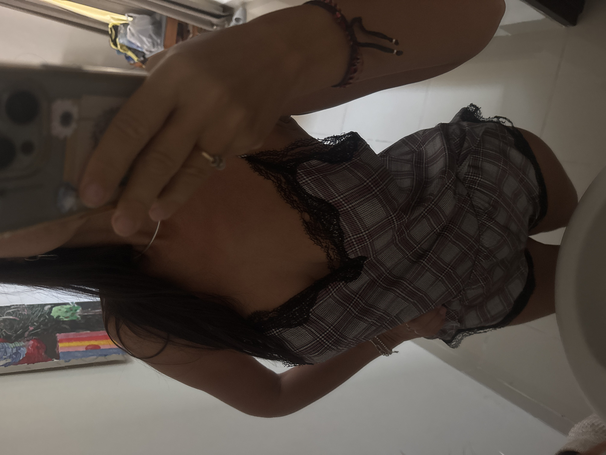 A medium shot shows a person in a bathroom taking a picture of themself. The person is wearing a patterned pajama top trimmed with black lace, and a black and red bracelet. A hand holds a phone to take the picture. In the background is a white sink and a painting. The location is Unknown, Unknown.