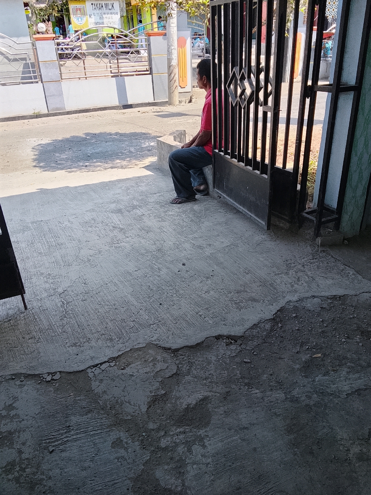 A person wearing a pink shirt and blue jeans is sitting on a concrete ledge next to a black metal gate. The gate is partially open, revealing a building and trees in the background. The ground in front of the gate is concrete and appears to be cracked and worn. The scene is outdoors, with sunlight casting shadows on the ground. The location is identified as Tayem, Indonesia.