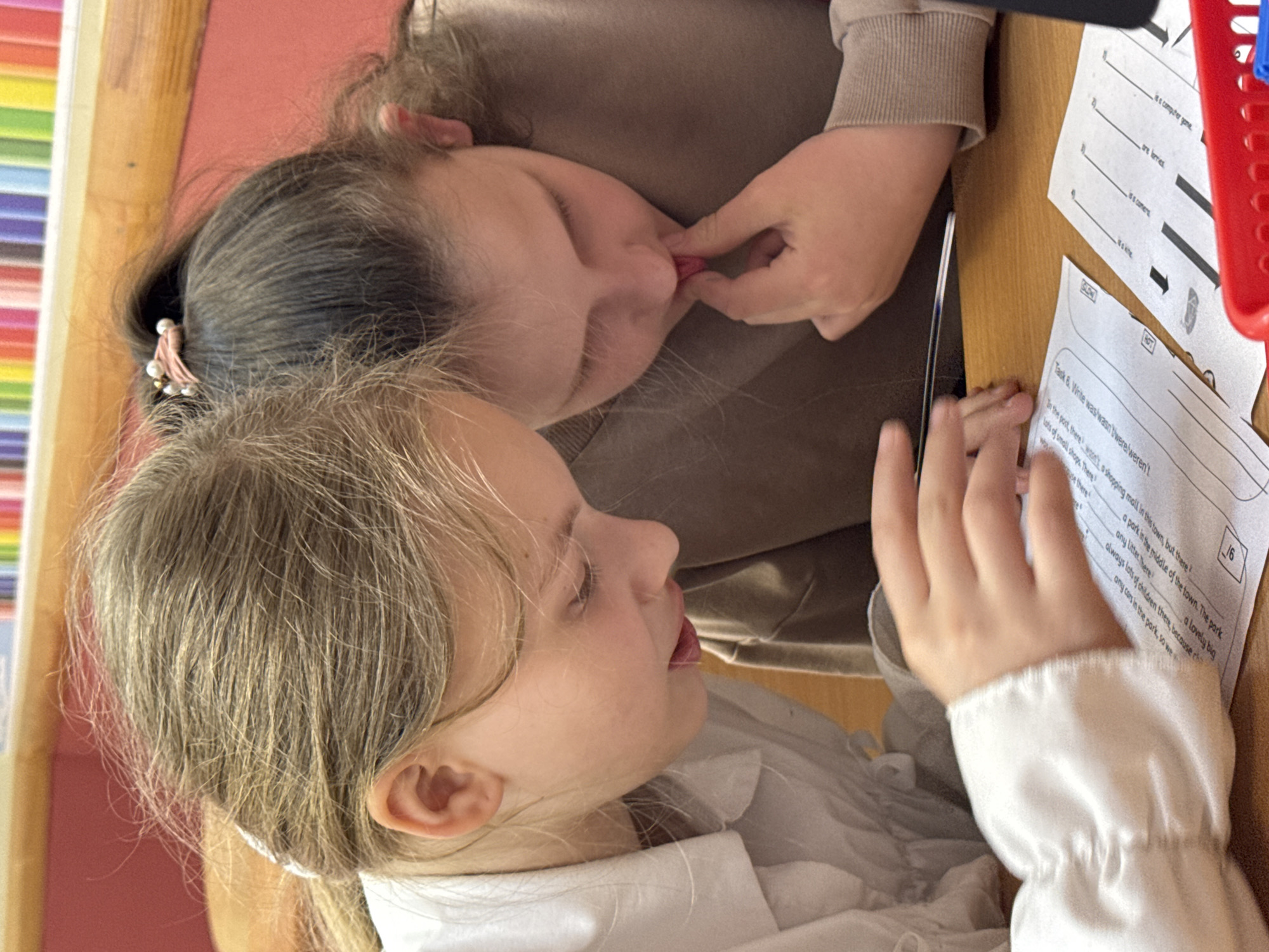 Two young girls are closely seated together at a wooden desk. One girl, with dark hair pulled back with a hair accessory, rests her face near the other girl's face and is holding the other girl's lip. The second girl, with light blonde hair, looks at a worksheet. A pencil lies on the desk beside the worksheet. The worksheet has a fill-in-the-blank task relating to grammar rules (was/wasn't/were/weren't). A red basket sits partly in view. There are light-colored bars in the background. The location of the city of Perm, Russia, is not discernible in the image.