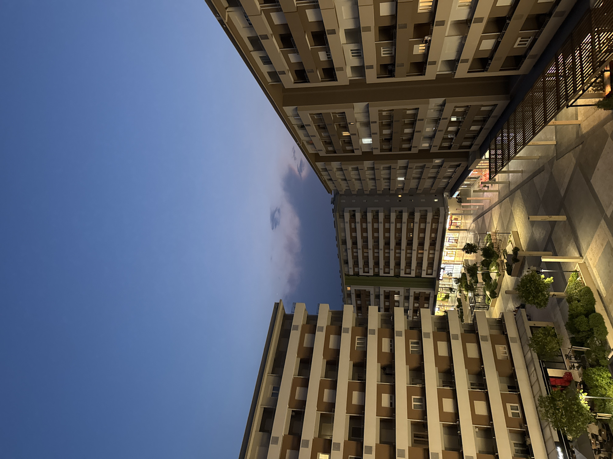 This is a high-angle medium shot of a modern urban development at dusk, featuring several multi-story apartment buildings. The sky is a deep, clear blue with a few wispy clouds. The buildings are characterized by their repetitive balconies and uniformly colored facades, predominantly beige and white. Several windows are illuminated, indicating occupancy. In the foreground, an open-air plaza is visible, with paved ground, planters containing small trees and shrubs, and outdoor seating areas for cafes or restaurants. Lights are on in ground-floor commercial spaces, and streetlights cast pools of warm light on the plaza. There are a few figures walking in the plaza, but they are small and indistinct. The overall impression is one of a contemporary, well-maintained residential and commercial area.