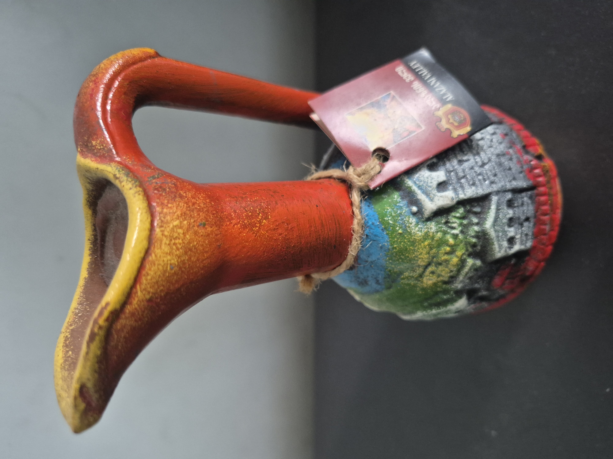 A colorful, handmade ceramic pitcher is displayed horizontally against a plain, out-of-focus grey background. The object's prominent handle and long spout are painted in vivid shades of red and yellow, featuring a speckled or distressed finish that adds to its rustic charm. A piece of rustic twine is tied around the neck of the pitcher, securing a small, reddish-brown rectangular tag.
The body of the pitcher, below the handle, is adorned with textured relief work, depicting architectural elements or a stylized cityscape in shades of blue, green, yellow, and grey, transitioning to a solid red base. No activity is taking place; the image is a still-life representation of the object.
Notable details include the tag, which features a logo resembling a coat of arms or seal and partially visible white text. Clearly discernible text includes "ALAZANI VALLEY" and "GEORGIA," indicating the object's origin. Additional numbers such as "9096 3550" are also visible.
The pitcher's vibrant colors and unique design suggest it is a decorative or souvenir item, originating from the Alazani Valley in Georgia. This media file was captured in Yerevan, Armenia.