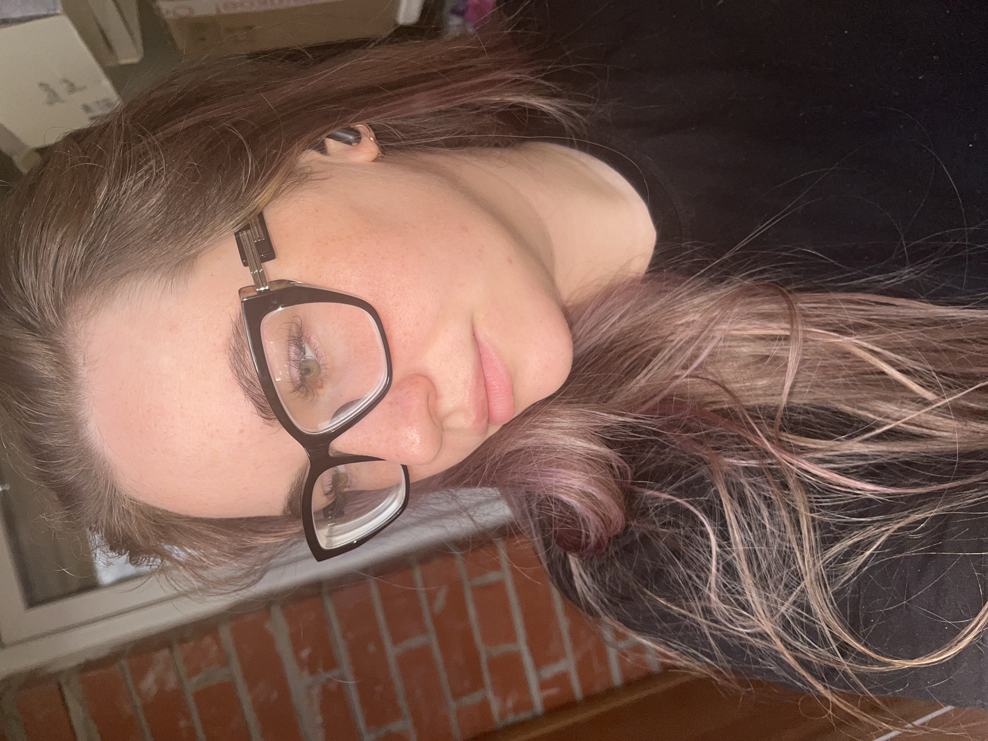 The image is a close-up, rotated portrait of a young person, likely female, with fair skin, freckles on the nose and cheeks, and green or hazel eyes. They wear black-framed glasses and a small dark earring or earbud in their left ear. Their lips are a natural pink, and their expression is soft and relaxed.
The subject's long hair, appearing as a blend of light brown or blonde with faded pink or purple undertones, cascades around their head. They are positioned as if lying down or leaning back against a dark, soft surface, possibly a piece of clothing or bedding.
In the background, visible to the bottom and left of the rotated image, is a section of a window frame and a reddish-brown brick wall, suggesting a setting indoors near a window or outdoors against a building. The upper and right parts of the background are dark, likely consisting of the soft material the person is resting against, with some indistinct, blurry objects further in the distance.
The lighting is even and soft, indicating indoor or subdued conditions. No specific activity is taking place beyond posing for the portrait. There is no visible text within the image. The provided location context is Chaikovsky, Russia, though this detail is not visually depicted in the photograph.