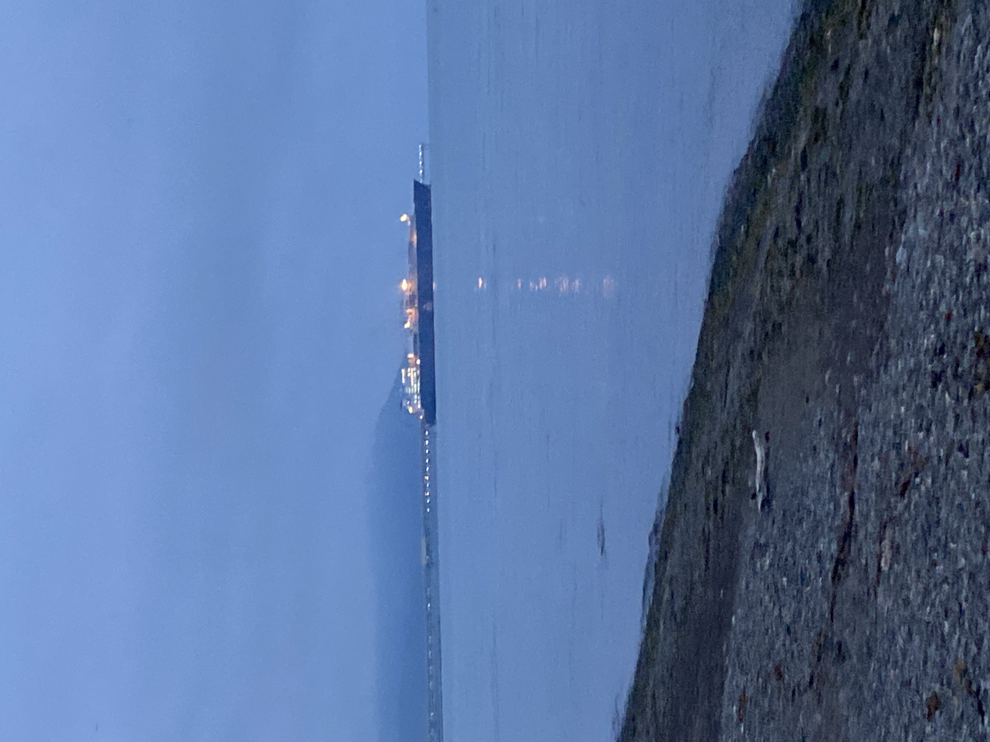 A wide shot captures a cargo ship illuminated by lights on the water in Korsakov, Russia. The ship is positioned in the mid-ground, with its hull and superstructure clearly visible. Lights are also visible along a distant coastline, suggesting a populated area. The foreground features a rocky shoreline. The sky is overcast, and the overall lighting indicates it is either dawn or dusk.