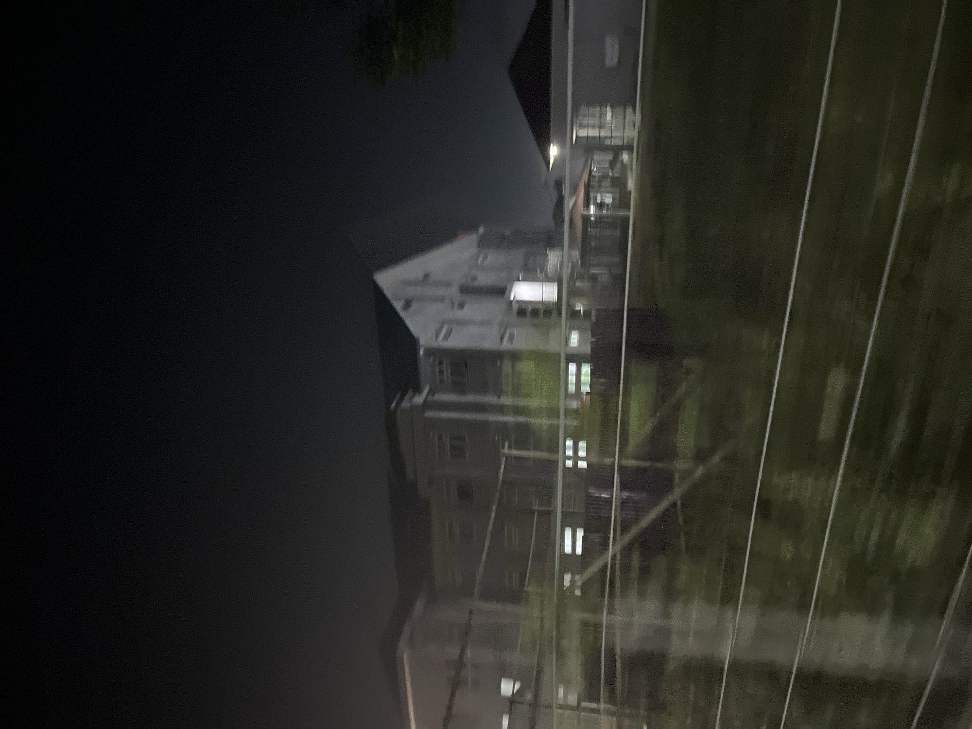 The image depicts a nighttime scene of a building complex at an institution named Kesehatonbandar in Indonesia. The structures are dimly lit, suggesting it is either night or very early morning. The main building appears to be a multi-story facility with windows that are partially illuminated. In the foreground, there are horizontal lines that might be part of a fence or railing, and beyond that, a dark, grassy area. The overall impression is one of quietude and potentially limited activity due to the low light conditions. The architecture of the buildings suggests an institutional setting, possibly a hospital, school, or research facility, consistent with the name "Kesehatonbandar" which implies a health-related or medical center.