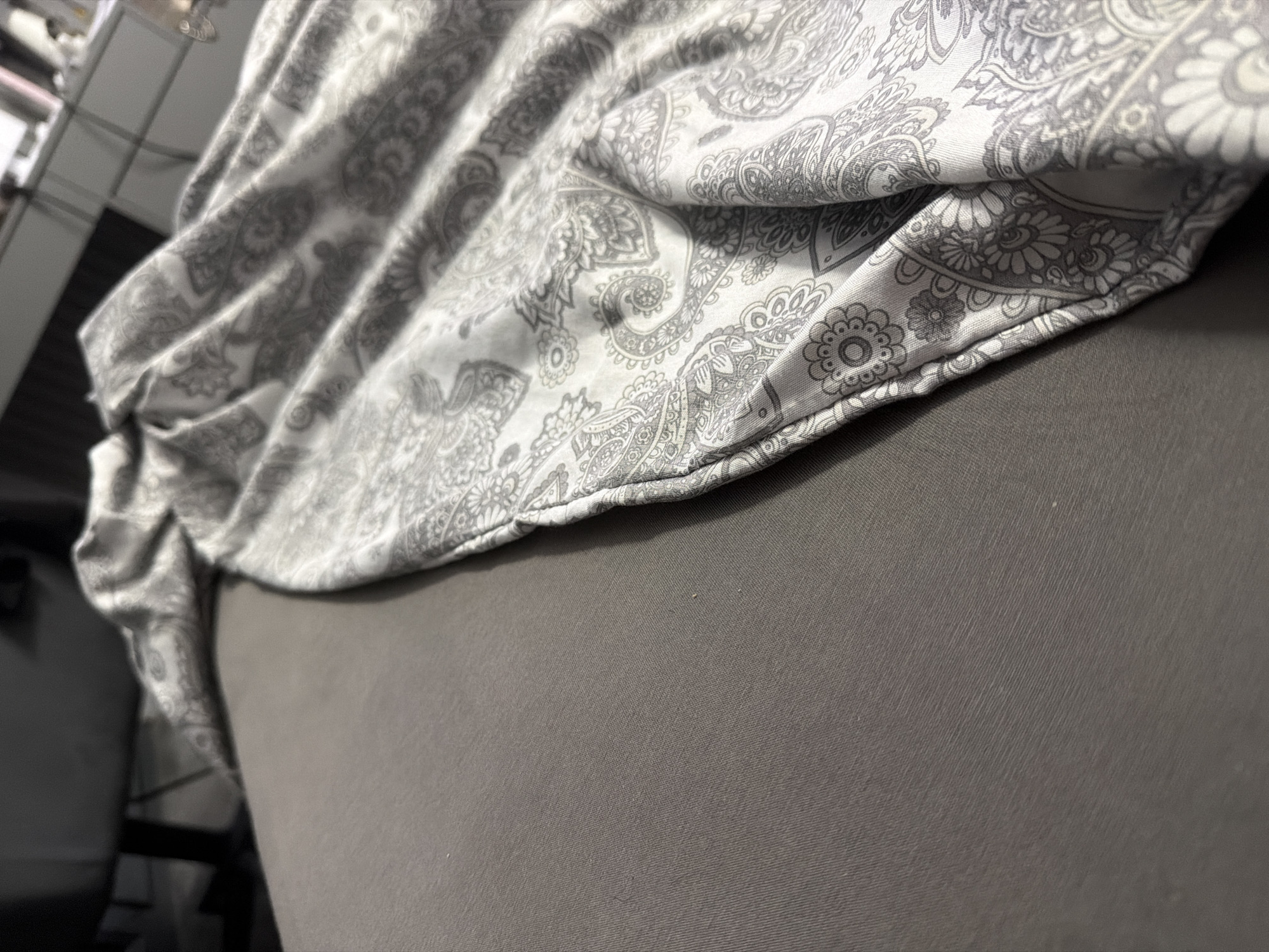 The image depicts a close-up of a gray textured surface, likely a bedsheet, with a folded section of fabric featuring a paisley pattern in shades of gray and white. The paisley pattern is detailed and repetitive. The lighting suggests an indoor setting, possibly a bedroom. There is no identifiable location in the city of Ruse, Bulgaria, or any indication of human presence or activity within the frame.