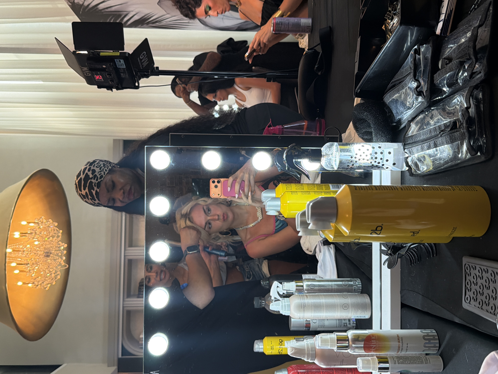 Here is a forensic description of the image:
**Scene:** The image appears to be taken backstage at an event or photoshoot, likely in Miami Beach, United States. This is suggested by the presence of a vanity mirror with bright lights, various makeup and hair products, and the general setup of a dressing room or preparation area.
**People:** Several individuals are visible, primarily in reflections in the mirror.
* A woman with blonde hair is holding a smartphone and taking a selfie. Her reflection is prominent in the center of the mirror.
* A makeup artist or stylist wearing a bandana is visible, possibly attending to the blonde woman's hair or makeup.
* Other individuals are visible in the background, some of whom seem to be working on hair or makeup.
**Objects:**
* A vanity mirror with multiple light bulbs framing its edges.
* Various bottles and containers of hair and makeup products are arranged on the counter in front of the mirror.
* A professional video light with the brand name "GVM" is visible.
* Clear makeup bags are visible.
* A gold-colored chandelier is partially visible in the background.
**Actions:**
* The blonde woman is taking a selfie.
* The makeup artist is presumably working on the blonde woman's hair or makeup.
* Other individuals in the background are engaged in similar preparation activities.
**Interactions:** The primary interaction is between the blonde woman and her makeup artist or stylist. Other interactions are suggested by the busy atmosphere of the backstage area.
**Environment:** The environment is cluttered with equipment and supplies, characteristic of a backstage or preparation area. The presence of a professional video light suggests that filming or photography is taking place. The high-end products and well-lit vanity indicate a professional setting.