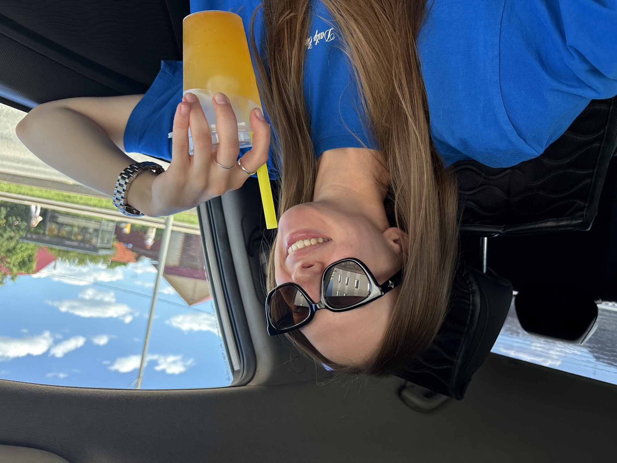The image shows a young woman in a car, holding a drink with a yellow straw and wearing a blue t-shirt. She has long brown hair and is wearing large black sunglasses. Her left arm is resting on the car door, and she is wearing a silver ring and a silver watch. The background shows a suburban street with houses and trees, under a blue sky with white clouds. The scene appears to be a sunny day, and the woman seems to be enjoying her drink. No specific landmarks or indicators that would pinpoint the city of Perm, Russia, are visible in the image.