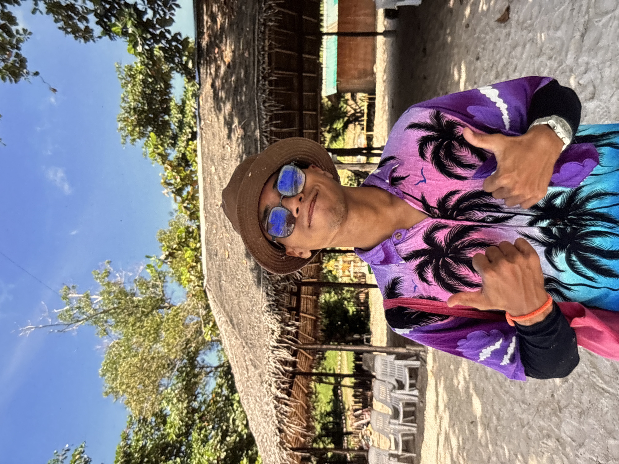 A medium shot captures a young person standing on a sandy beach in Chalong, Thailand. They are wearing a brown bucket hat, sunglasses with blue mirrored lenses, and a vibrant, colorful button-up shirt featuring a tropical palm tree and bird print with a gradient from purple to pink to blue. The person is wearing a black long-sleeved shirt underneath the button-up and a pink bag is slung over their left shoulder. They are giving two thumbs up with their hands. In the background, a thatched-roof hut with wooden posts is visible, and rows of white plastic chairs are arranged on the sand in front of it. Lush green trees with broad leaves and a bright blue sky with a few wispy clouds complete the tropical beach scene.
