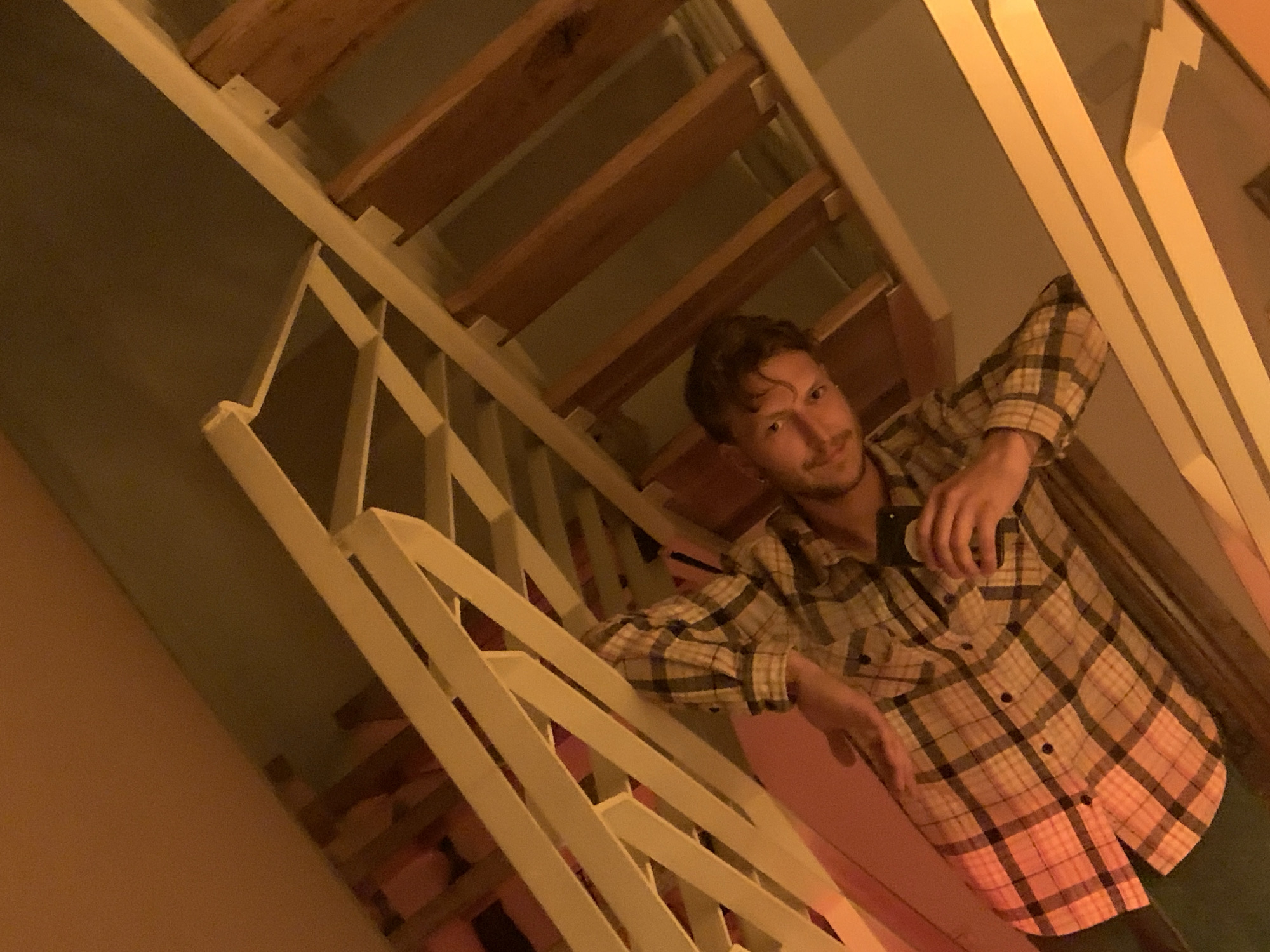 A man with short, curly brown hair and a light beard is leaning on a white stair railing in what appears to be an interior setting. He is wearing a long-sleeved, plaid button-down shirt with a combination of tan, brown, and red hues. He seems to be holding a phone in his right hand, possibly taking a photo. The stairs behind him are constructed of wooden planks. The lighting in the scene is warm, casting a yellow hue throughout. Based on the image content, there is no information about the location being in Tel-Aviv, Israel.