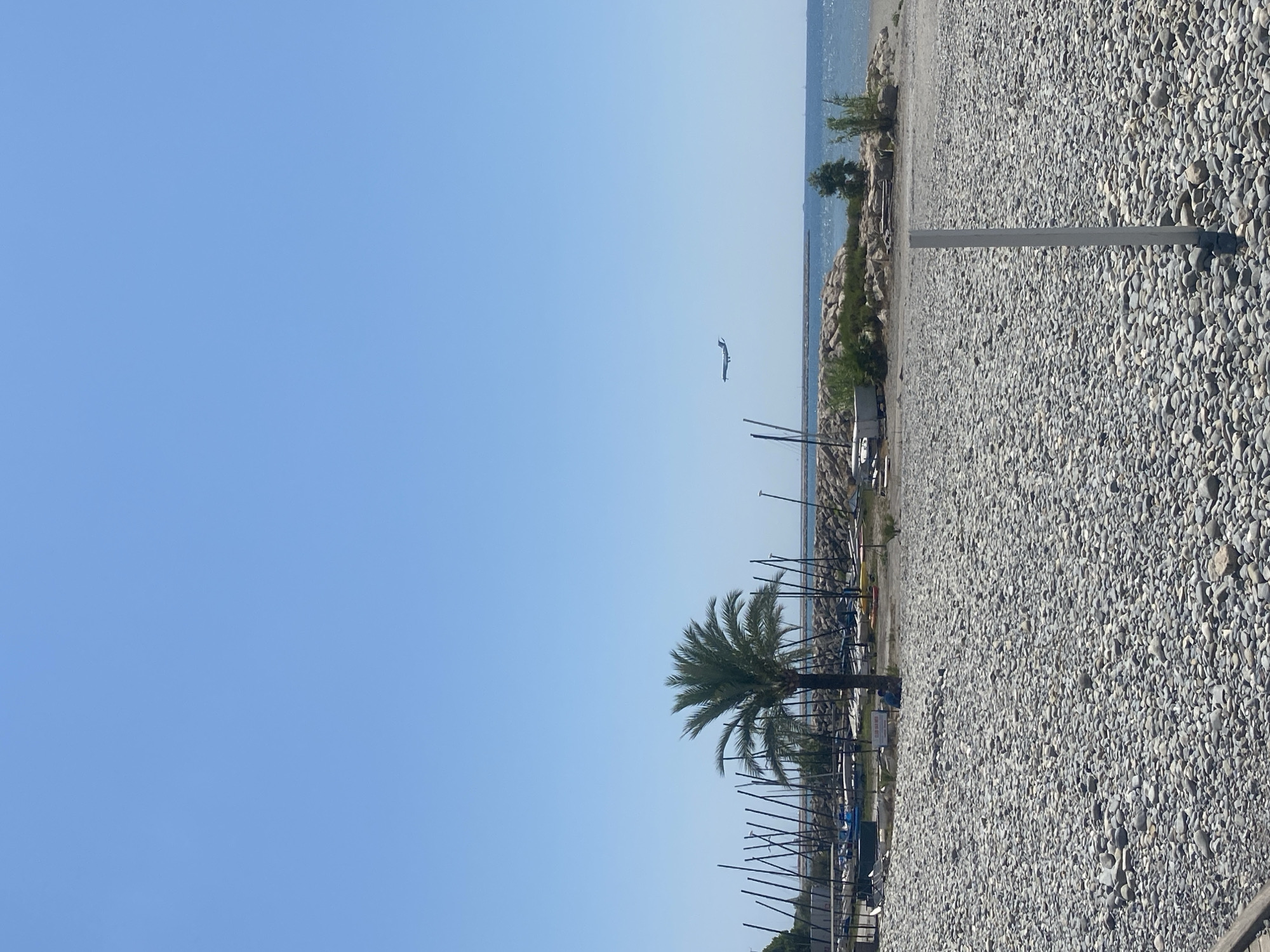 This is a vertical, medium-shot, outdoor photograph taken on a clear, sunny day in Saint-Laurent-du-Var, France. The foreground is dominated by a densely packed, grey pebble beach, with a light grey vertical pole in the lower right quadrant. In the mid-ground, beyond the beach, there is a harbor or marina area with numerous sailboat masts, a palm tree, some small boats on trailers, and a rocky breakwater extending into the bright blue sea. A small airplane is visible in the sky, flying horizontally from left to right. The sky is clear and blue, with no clouds. The scene depicts a coastal environment, likely a beach or harbor area in a French Riviera town.
