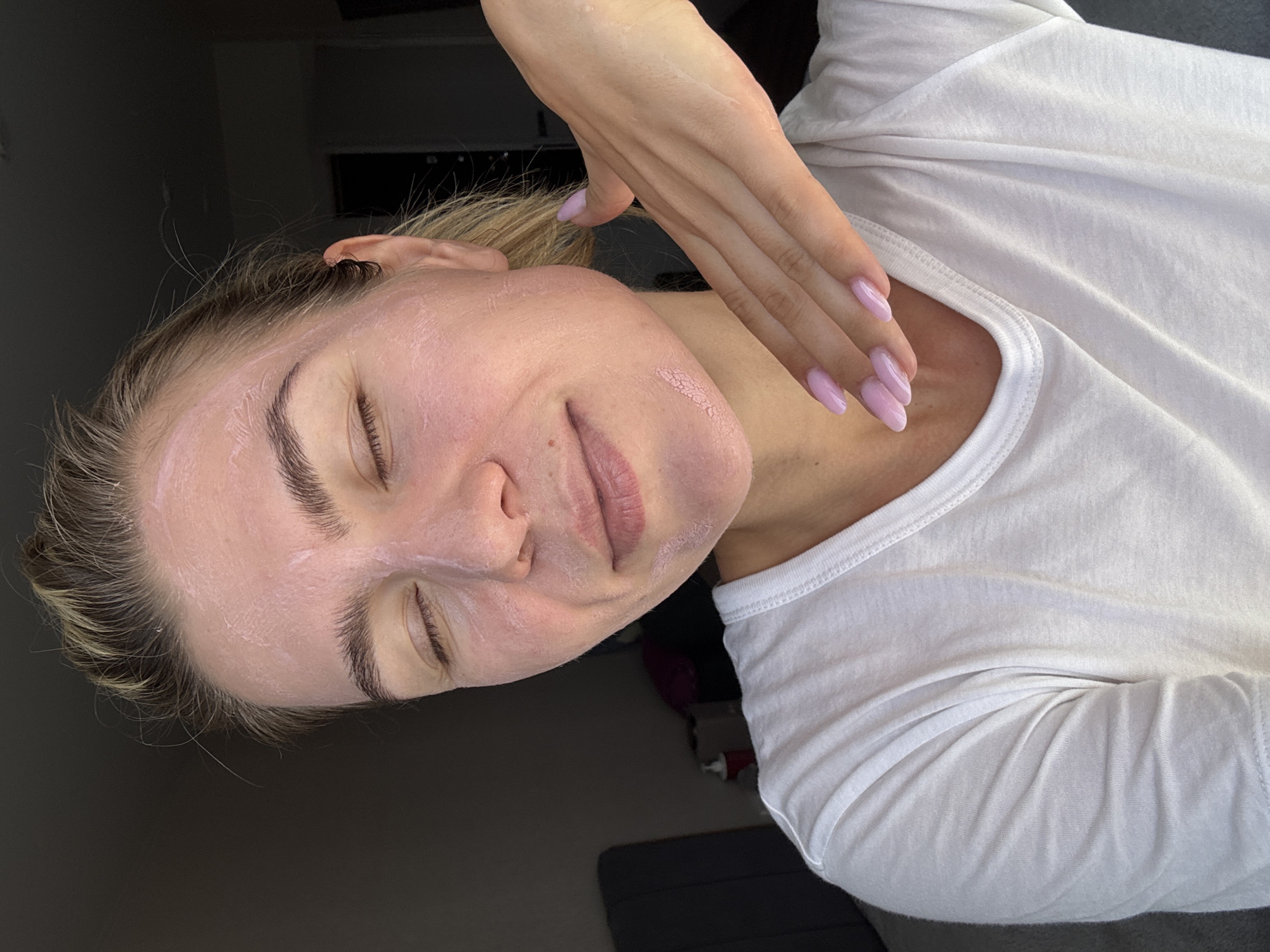 A young Caucasian woman with blonde hair pulled back is shown from the chest up, indoors. She is wearing a white t-shirt and has a pink facial mask applied to her face. Her eyes are closed, and she is smiling slightly. Her right hand is raised to her jawline, with her fingers lightly touching her face. The woman's fingernails are painted a light pink color. The background is dimly lit, with a dark wall and a hint of a sofa visible on the left. The lighting suggests it is daytime, possibly near a window.