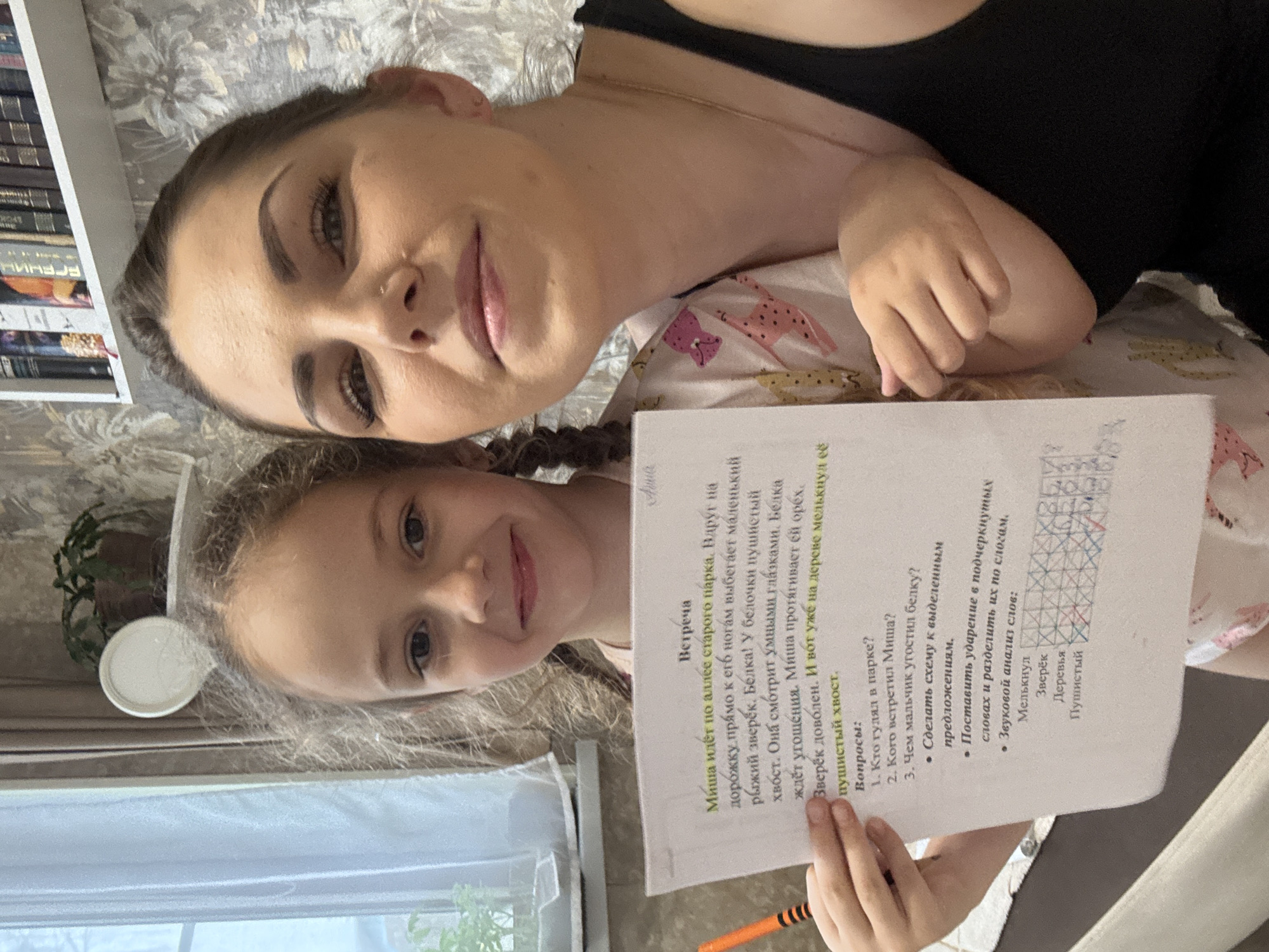 Here's a detailed description of the image:
**Scene:** A woman and a young girl are posing for a selfie, holding a printed page with a children's story and related exercises.
**People:**
* A young woman with dark hair and fair skin.
* A young girl with blonde hair styled in braids.
**Subjects:**
* The printed page contains a children's story titled "Встреча" (Meeting) and exercises related to the story.
**Objects:**
* The printed page
* An orange pen
* Bookshelf in the background
* Window
**Actions/Interactions:**
* The woman is holding the phone or camera to take the selfie.
* The girl is holding the printed page, possibly pointing at it with a pen.
* Both are looking at the camera, smiling.
**Environment:**
* The setting appears to be indoors.
* The background shows a bookshelf, a wallpapered wall and a window.
**Location:** Based on the OCR text and the name given, the location is Razdolie, Russia.