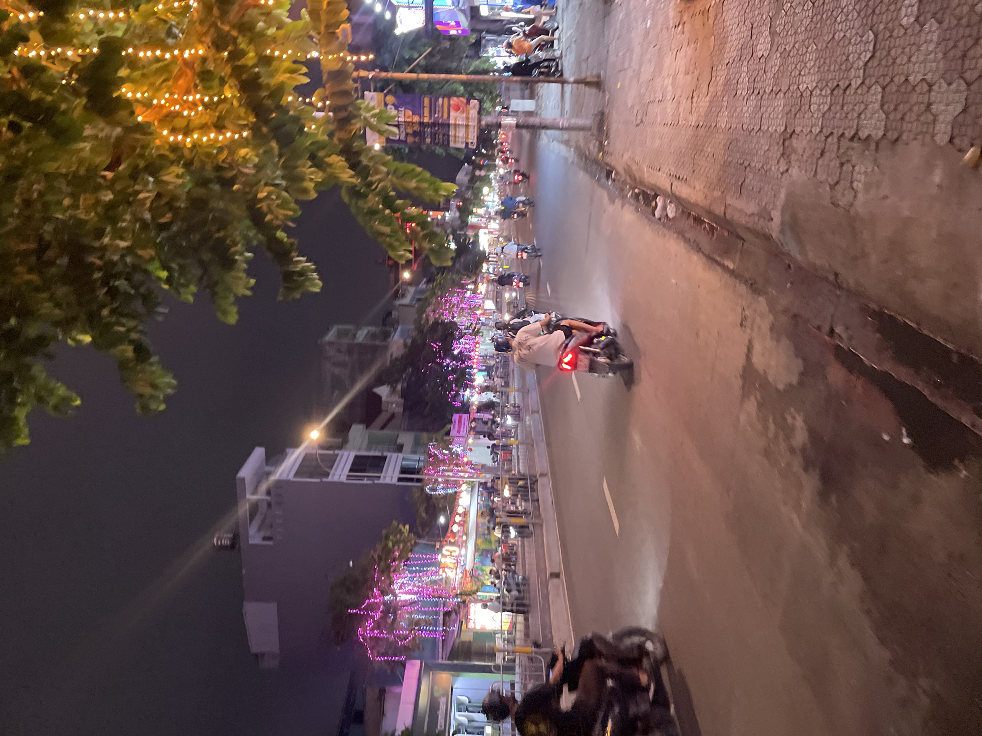 This is a medium-angle, nighttime photograph of a busy street in Thuận An, Vietnam, likely a commercial or entertainment district, as indicated by the abundance of decorative lights. The foreground shows a paved road with a slight sheen, suggesting recent rain or humidity. A dark-colored motorcycle with a rider wearing a dark shirt and helmet is moving away from the viewer on the left side of the road.
The middle ground is occupied by a multi-lane street with several motorcycles in motion. One motorcycle in the center has a rider wearing a light-colored shirt and helmet. On the right side of the road, there's a sidewalk paved with hexagonal tiles. Along the left side of the street, buildings are visible, adorned with numerous strings of pink and purple lights, creating a festive atmosphere. Some of these buildings appear to be businesses, possibly restaurants or shops, with people seated outside.
In the background, more buildings rise, some illuminated with lights. A tree with foliage and strings of warm-toned decorative lights hangs over the right side of the scene, partially obscuring the sky. The overall impression is a lively urban environment at night, characterized by traffic and vibrant lighting.