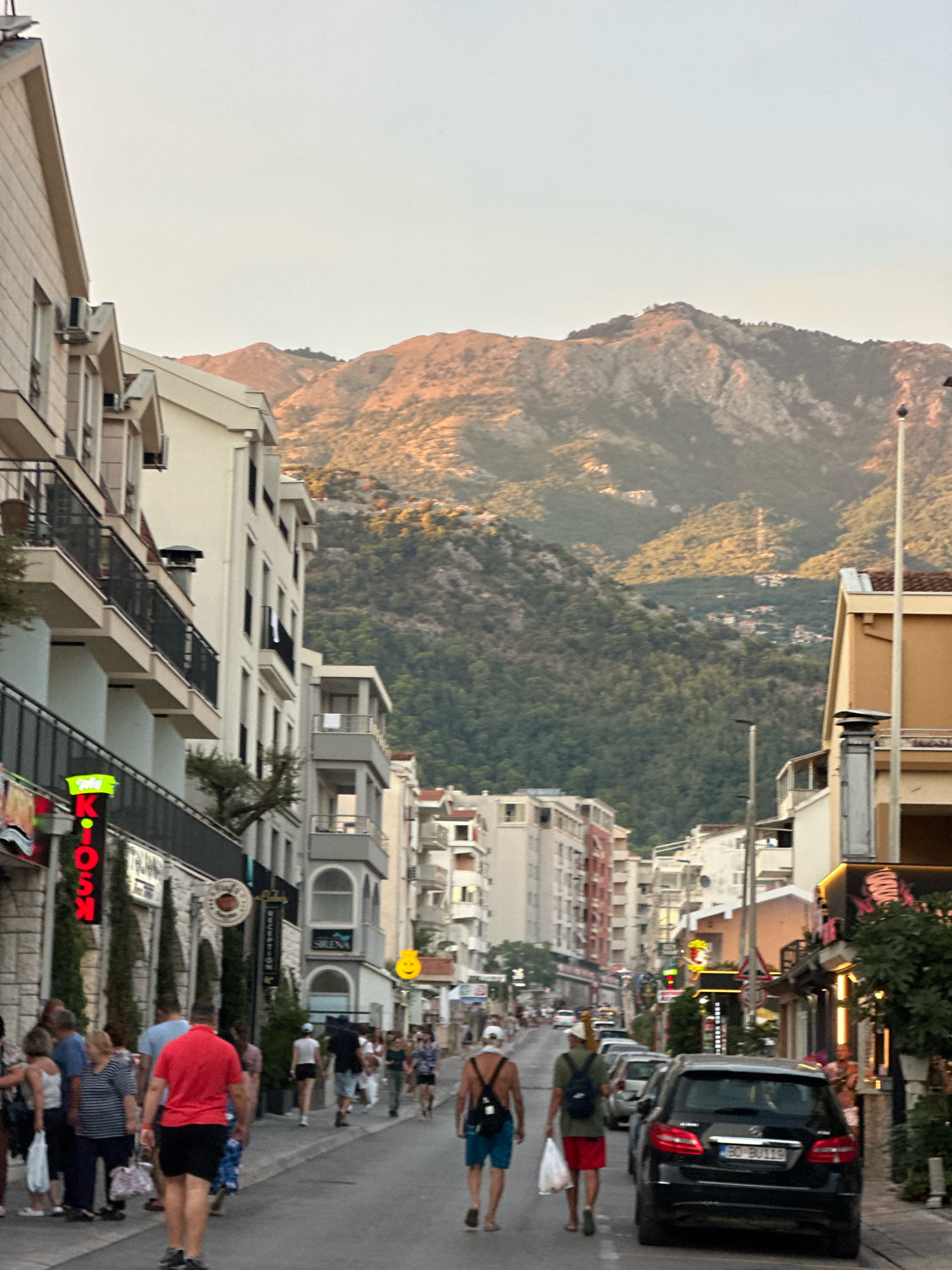 This is a vertical, full-shot, eye-level, outdoor photograph of a street scene in Budva, Montenegro, during the golden hour. The street is lined with multi-story buildings on both sides, with shops and businesses on the ground floor. The buildings have a mix of modern and traditional architecture, with balconies and awnings. In the background, a large, tree-covered mountain range dominates the skyline, with its peaks illuminated by the setting sun.
Several people are walking along the street, some in groups and some alone. They are dressed in casual summer clothing, suggesting a warm climate. There are also several cars parked along the street and some in motion. One black Mercedes-Benz is prominent in the foreground. The street is paved and appears to be a pedestrian-friendly area with shops and restaurants. The lighting suggests late afternoon or early evening, with a warm, soft glow. A neon sign with the word "KIOSK" in red lettering is visible on the left side of the street, and a yellow smiley face icon is also visible on a sign. The overall atmosphere is one of a vibrant, bustling street scene in a popular tourist destination.