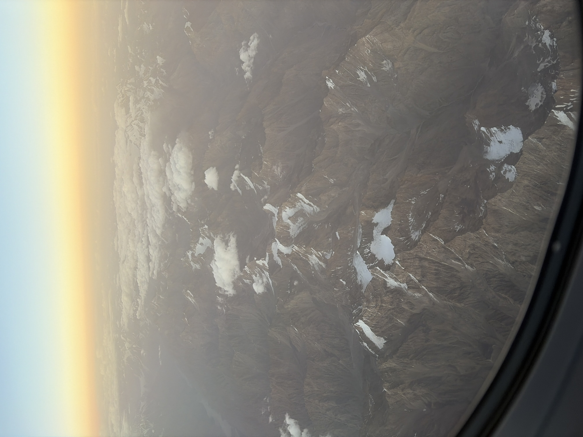 An aerial view captures the rugged, snow-capped peaks of the Pamir Mountains in Tajikistan, as seen from an airplane window. The landscape is dominated by sharp, rocky ridges with patches of white snow clinging to their upper slopes. Valleys and ravines carve through the mountains, creating a stark and dramatic topography. Above the mountains, a layer of clouds partially obscures the view, with a hazy, warm-toned sky suggesting either sunrise or sunset. The lower right corner of the image shows the dark, curved frame of the airplane window, indicating the perspective is from inside the aircraft. The scene is one of vast, remote wilderness, characterized by its mountainous terrain and atmospheric conditions. The exact location within Vanj, Tajikistan, is not discernible from the visual information alone, but the geological features are consistent with the region's mountainous geography.