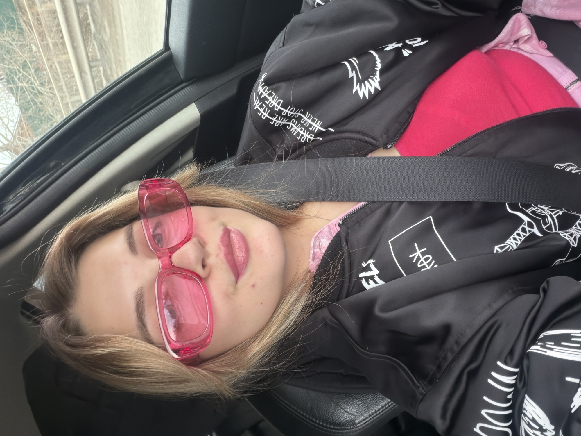 A young woman wearing pink sunglasses and a black jacket with white writing is sitting in the passenger seat of a car. She is wearing a seatbelt. A glimpse of the outside shows trees without leaves. The location is Больша́я Ши́льна, Russia.