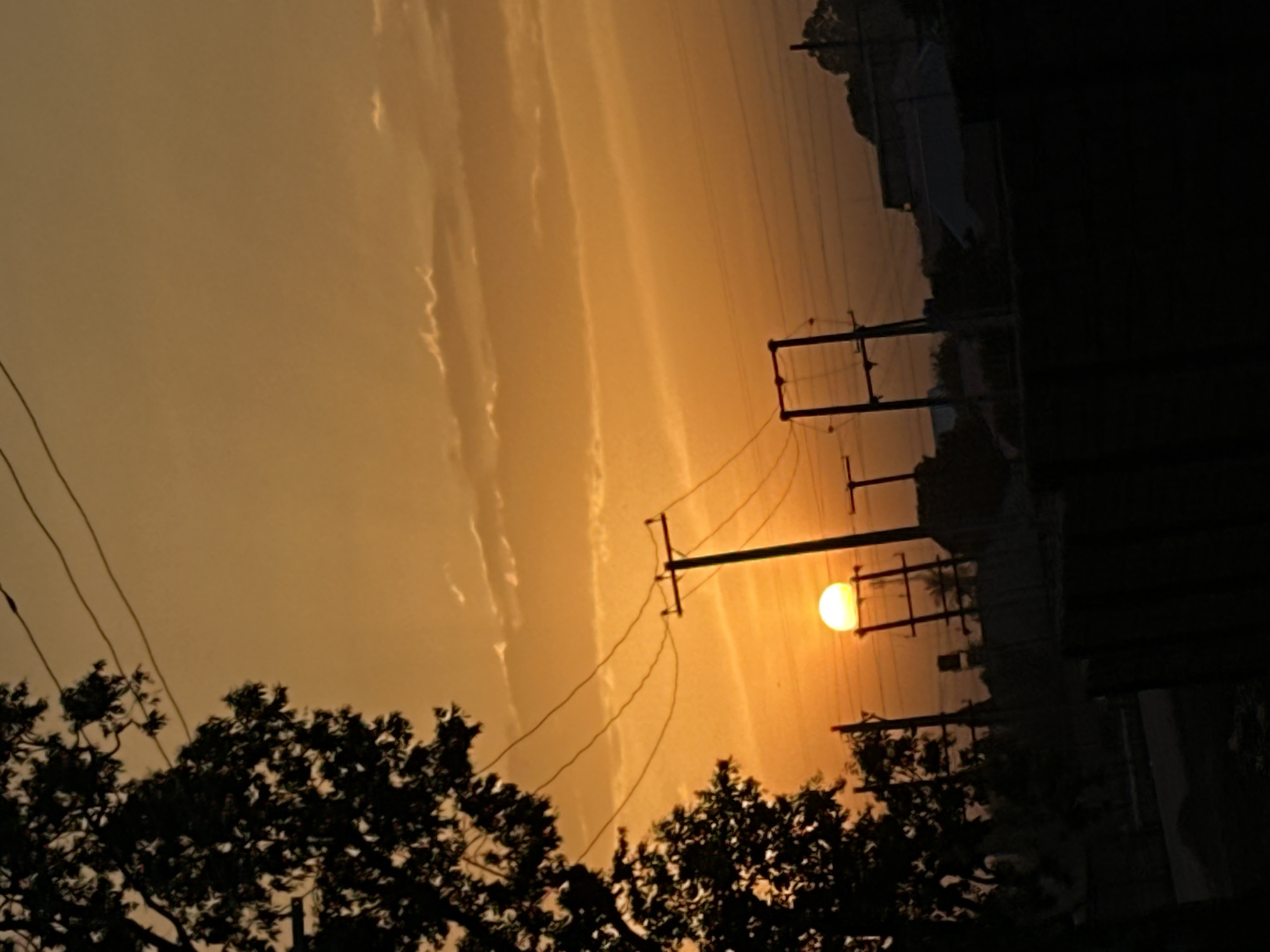 The image shows a sunset in Bukuru, Nigeria. The sky is orange and there are clouds in the sky. There are trees and power lines in the foreground. The sun is visible through the trees. Buildings are visible to the right of the power lines and trees. The overall scene depicts a tranquil sunset setting in an urban environment.
