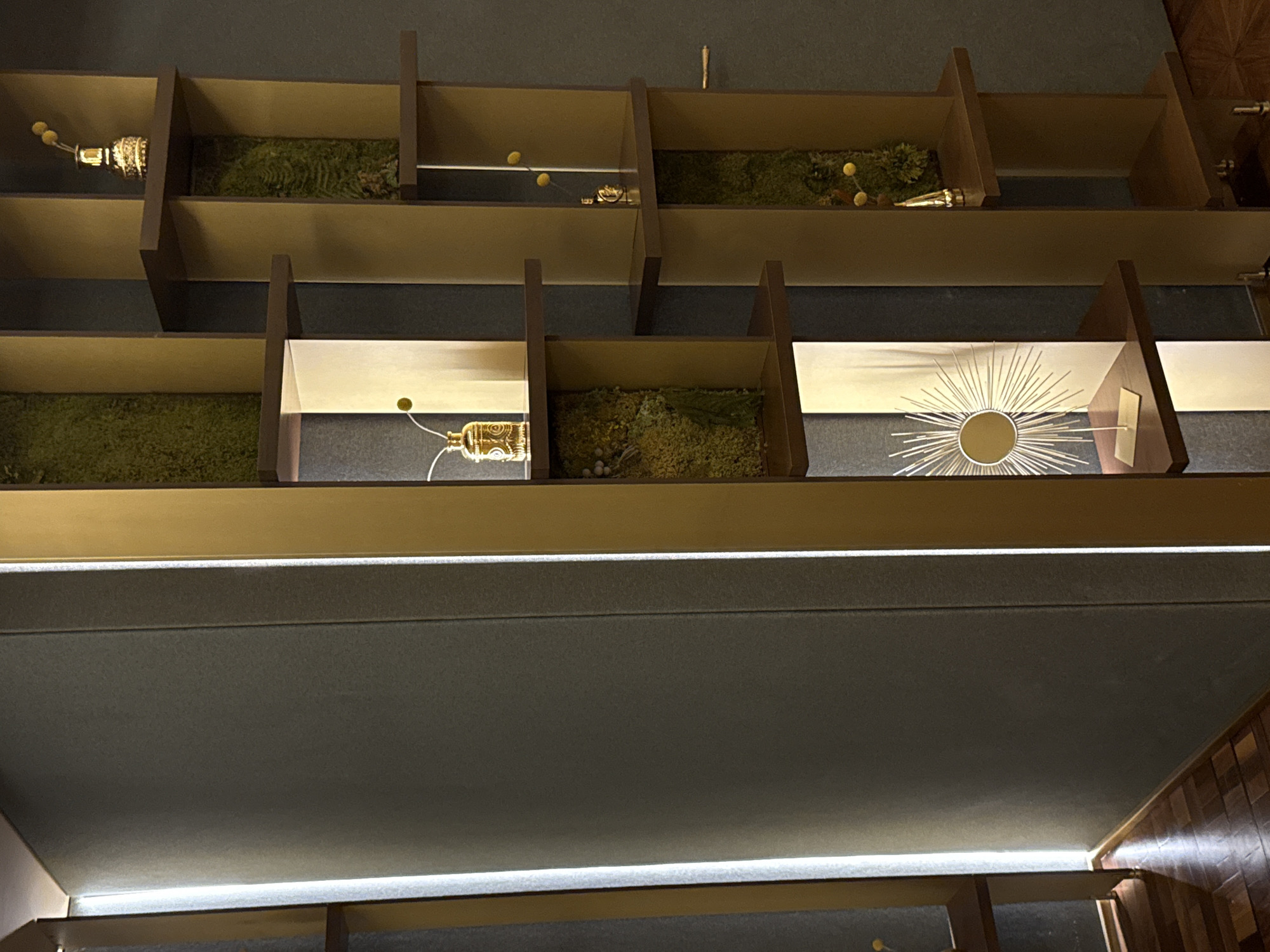 This media file depicts a series of display shelves, likely in a religious or commemorative setting, possibly in Trieste, Italy. The shelves are constructed with dark brown vertical dividers and golden horizontal surfaces. Each shelf contains a diorama-like arrangement, featuring green moss or similar material as a base.
Scattered within these mossy displays are various small, golden objects that appear to be religious artifacts or decorative elements. Some resemble small chalices or reliquaries, intricately detailed with patterns. In the middle row, on the rightmost shelf, a larger, ornate golden object with radiating rays, resembling a monstrance or sunburst, is prominently displayed against a white background. Small yellow spheres, possibly decorative baubles or symbolic elements, are visible in some of the upper shelves.
The lighting appears to be artificial, with some shelves illuminated from within or from above, casting soft light and shadows. The overall impression is one of quiet reverence and display. No people are visible, and the scene is static, with no specific activity taking place. The context of Trieste, Italy, suggests a cultural or historical significance to these displayed items, possibly within a church, museum, or a dedicated memorial space. The time of day and weather are not discernible from the image. There is no visible text.
