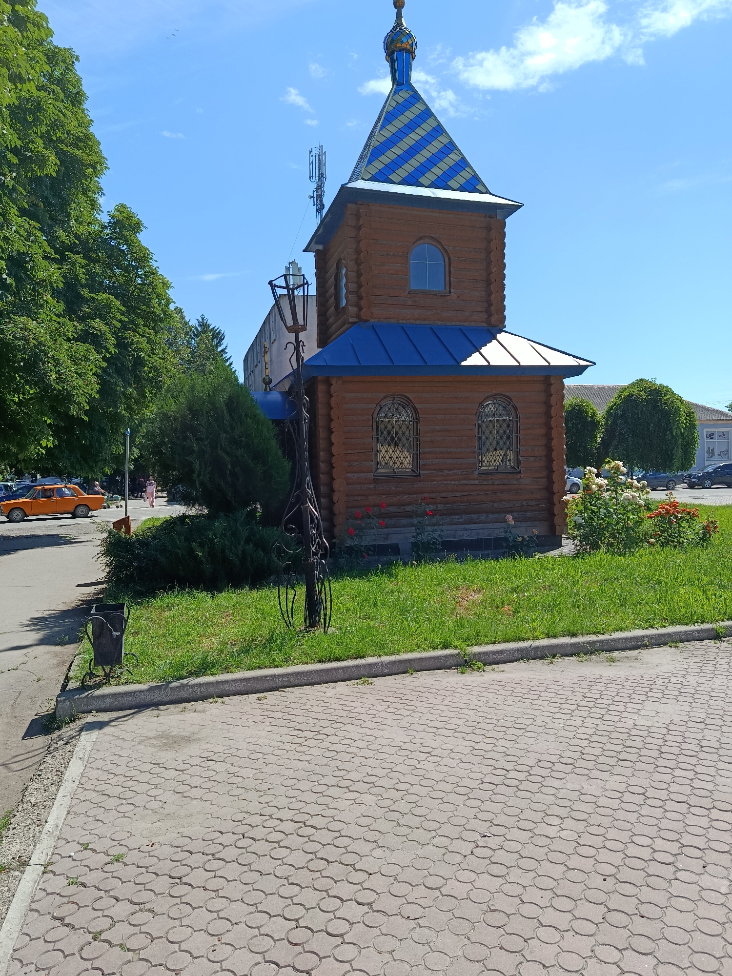 In the city of Yampil, Ukraine, a wooden chapel with a blue and yellow patterned roof stands on a grassy area adjacent to a paved walkway. The chapel features arched windows with decorative grilles and a conical blue roof topped with a golden spire. To the left of the chapel, a black metal lamppost with intricate scrollwork stands on the grass. Behind the chapel, a larger, gray building with windows is visible. A street runs in front of the chapel, with an orange car parked on the side. Several trees with green foliage are present, including a large one on the left and two topiaries to the right. In the background, more buildings and trees can be seen under a clear blue sky with a few white clouds. A trash receptacle is placed near the edge of the paved walkway.