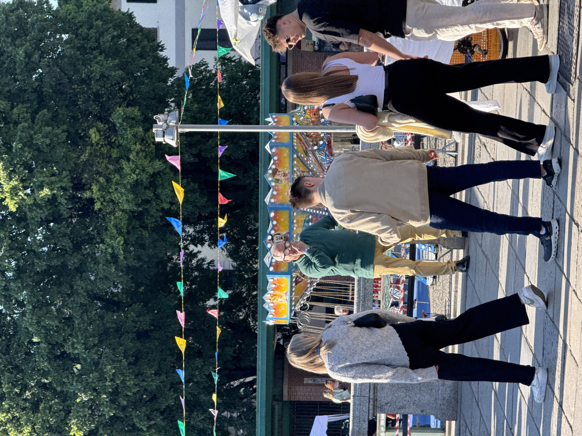 The image captures an outdoor urban scene in Milan, Italy, filled with people, colorful decorations, and a fairground attraction under bright daylight.
In the foreground, several individuals are visible, casually dressed and walking or standing on a paved area, casting long, distinct shadows, suggesting late afternoon sun. From left to right, a woman with blonde hair, seen from behind, wears a light grey textured cardigan and black pants. Next to her, a man in a dark green long-sleeved shirt and beige pants, wearing glasses, looks towards the right. Further right, another man in a beige jacket and dark blue pants walks. A woman with long brown hair, seen from behind, wears a white tank top and black pants, carrying a black shoulder bag. Behind her, a man in a black t-shirt and light-colored pants stands next to another person partially obscured.
The mid-ground features a vibrant carousel or fair ride, richly decorated with colorful panels. Visible details on the ride include a character with the number "80" and an image of a princess. Behind the ride, a red sign with the "pepsi" logo is faintly discernible.
The background on the left is dominated by lush green trees, likely part of a park or large avenue, with parts of a white building visible above them. A street light or traffic signal is also present. Strung across the scene at various heights are lines of colorful triangular bunting, adding to a festive atmosphere, possibly indicating a local fair, market, or celebration. The overall setting appears lively and open.