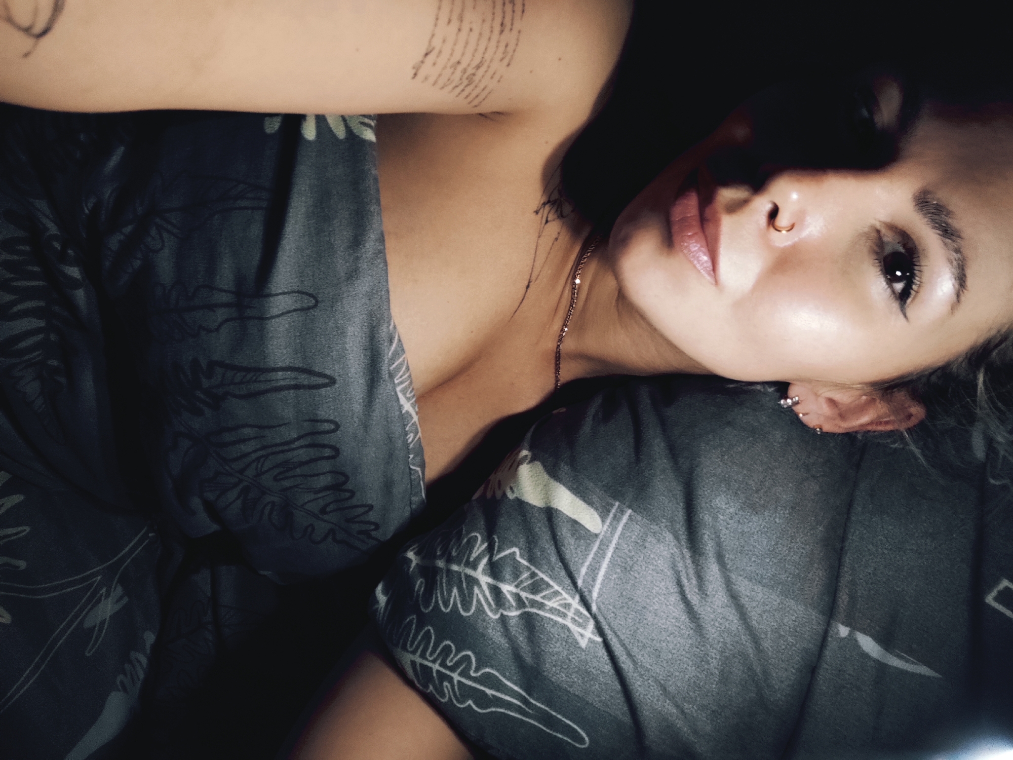 The image is a close-up, dimly lit, low-angle shot of a woman lying down, possibly in bed, based on the presence of patterned bedding. Her face is turned towards the camera, with a visible nose ring and multiple ear piercings. She has visible tattoos on her upper arm and neck. The bedding has a dark background and a lighter-colored leafy pattern. The location in Chaikovsky, Russia is not discernable from the provided image.