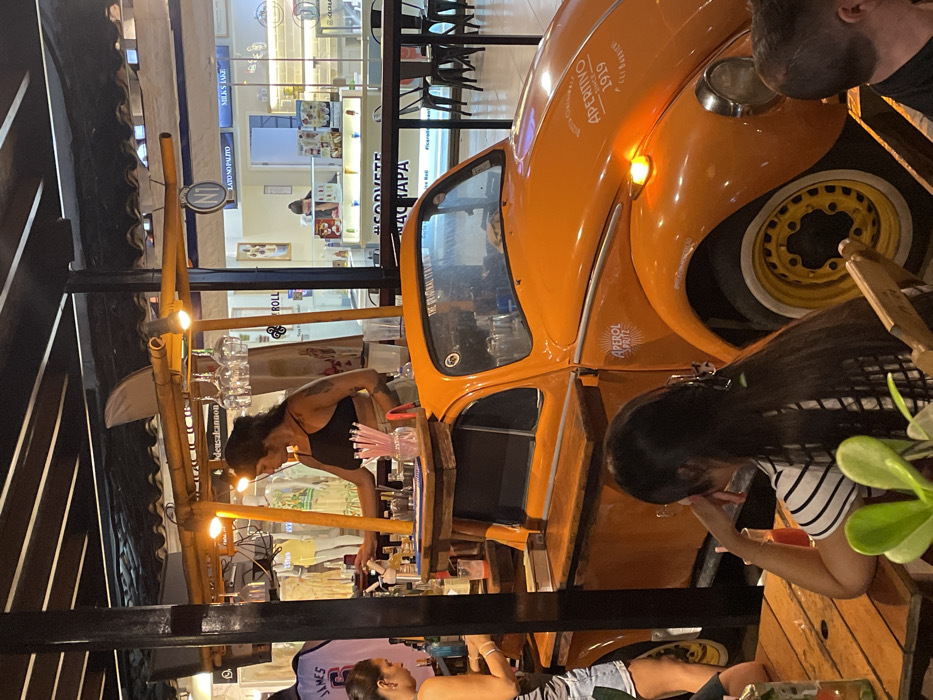 This image captures a nighttime scene within a commercial area of Armação Dos Búzios, Brazil, likely a pedestrian street or open-air food court. The central element is an orange Volkswagen Beetle, customized into a mobile bar. The Beetle displays "APEROL SPRITZ" branding on its front fender and side, and "APERITIVO 1919" on the hood, with matching yellow wheels. The rear portion of the vehicle is fitted with a wooden bar counter and an overhead wooden framework supporting warm light fixtures, from which several wine glasses hang. A white surfboard fin is attached to the top of this framework.
A female bartender with dark hair and arm tattoos, wearing a black bralette, is actively preparing drinks behind the bar, which holds various bottles, shakers, and a container of pink straws. To her left, another woman with dark hair, wearing a light-colored top and shorts, stands near the bar, appearing to engage with the service. In the foreground, a third woman with long dark hair, wearing a striped shirt, is seated at a wooden table, her left hand resting on her chin, looking towards the bar. A red drink is visible on the table. Partially visible in the bottom right corner is the head of a fourth individual, a male, also seated at the table.
In the background, several brightly lit commercial establishments are visible through glass storefronts. One prominent shop, likely an ice cream parlor, displays signs such as "#SORVETE" (Ice Cream) and "GELATO NO PALITO," with visible display cases of frozen desserts. Other shops feature retail merchandise, including a sports jersey marked "JAMES 6." The scene is illuminated by artificial lights from the bar and the surrounding businesses, under what appears to be a covered, open-air structure.