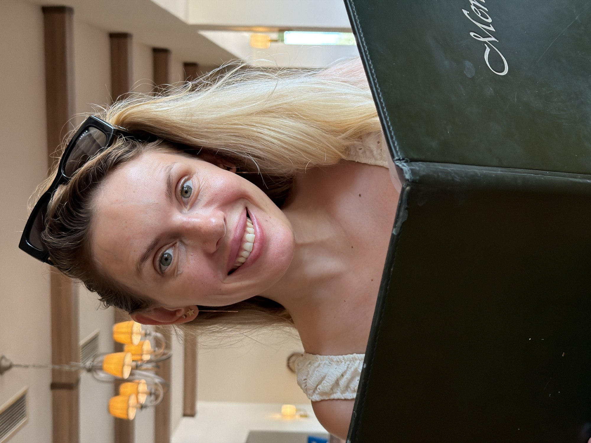 In Bigor, Montenegro, a woman with blonde hair and sunglasses perched on her head is smiling at the camera while holding a dark green menu. She is wearing a white top with ruffled details on the shoulders. The background suggests an indoor dining area with wooden beams and chandeliers, characteristic of a restaurant or hotel in the region.