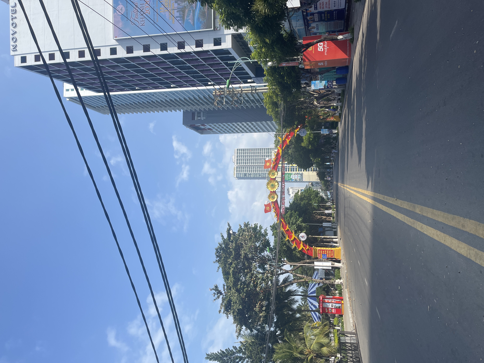 The image shows a wide, empty asphalt road in Nha Trang, Vietnam. The road has yellow double lines marking the center. An arched structure, decorated with red and yellow banners and sunflowers, spans the road. Tall buildings, including the Novotel, line the right side of the street, with several power lines crisscrossing the sky. Trees and palm trees line the left side of the road, with a few blue and white striped awnings visible. Red kiosks and large red advertising boards with "5G mobifone" are present on the sidewalk. The sky is blue with scattered white clouds. There are no people or vehicles visible on the road, suggesting it may be early morning or during a special event. The scene is characteristic of a coastal city in Vietnam, with a mix of modern high-rise buildings and tropical vegetation.